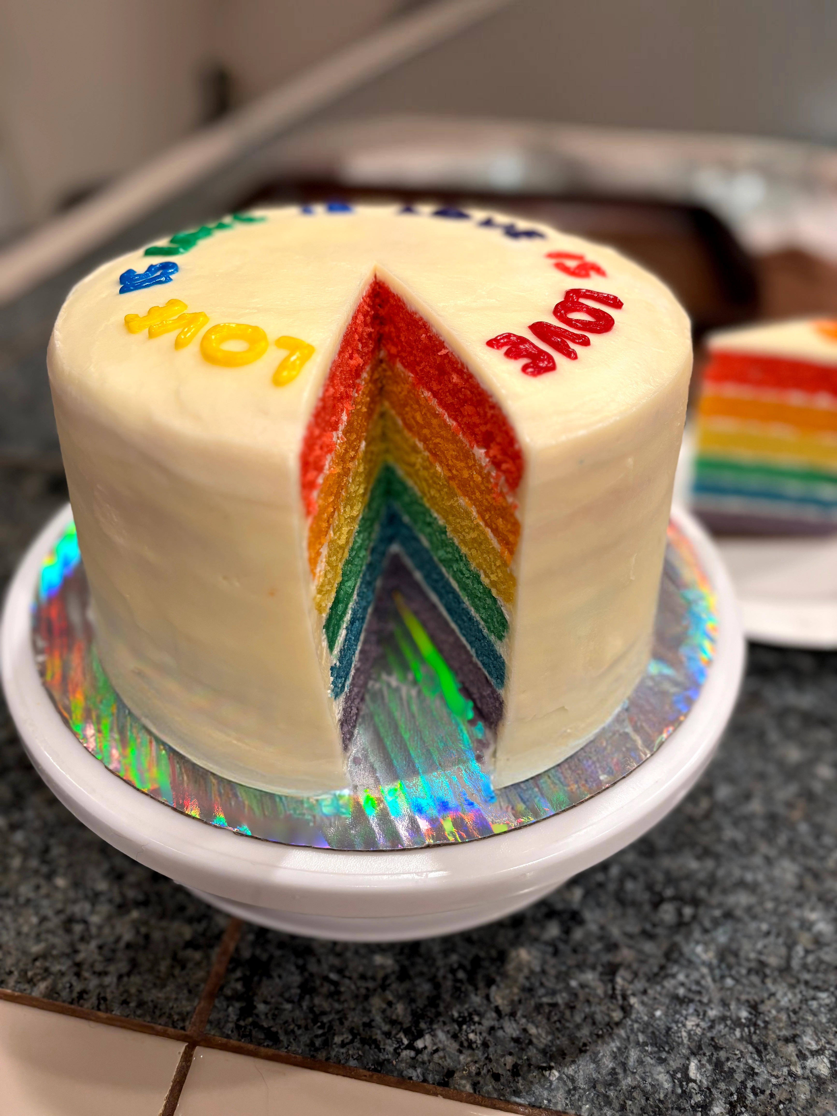 A round rainbow layered cake with white frosting, decorated with colorful words, on a white stand, with a slice removed showing rainbow layers.
