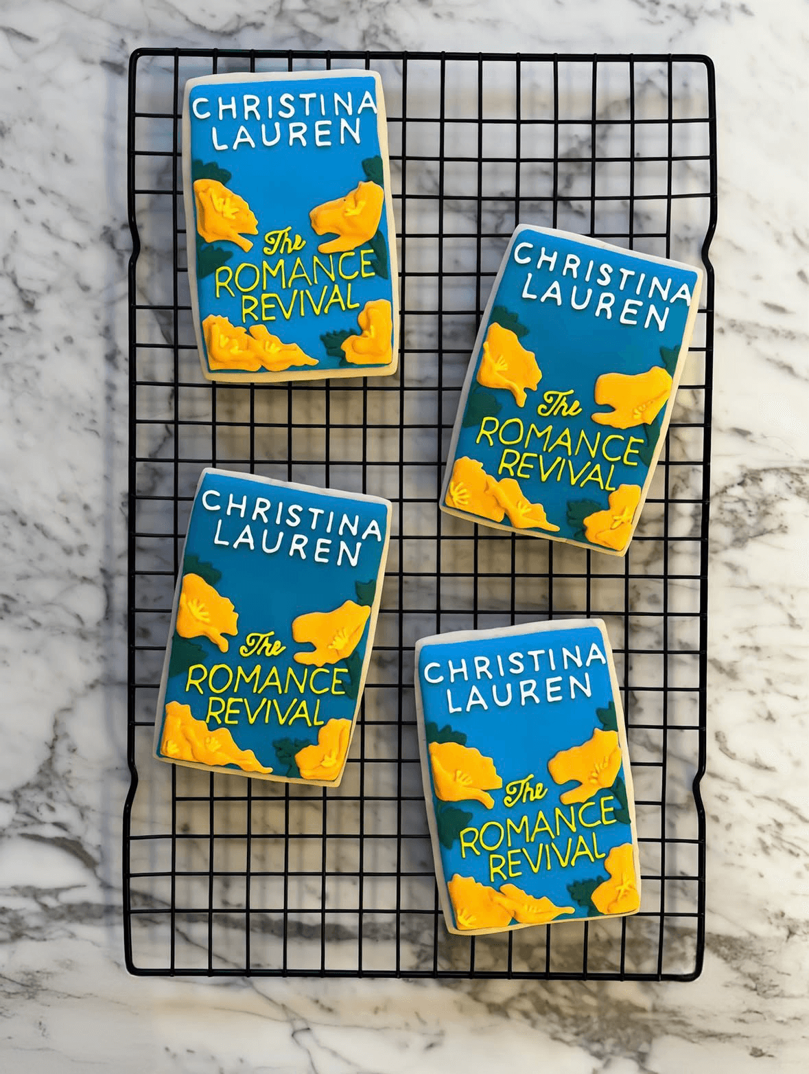 Four copies of a book titled 'The Romance Revival' by Christina Lauren on a wire rack, with a marble surface background.