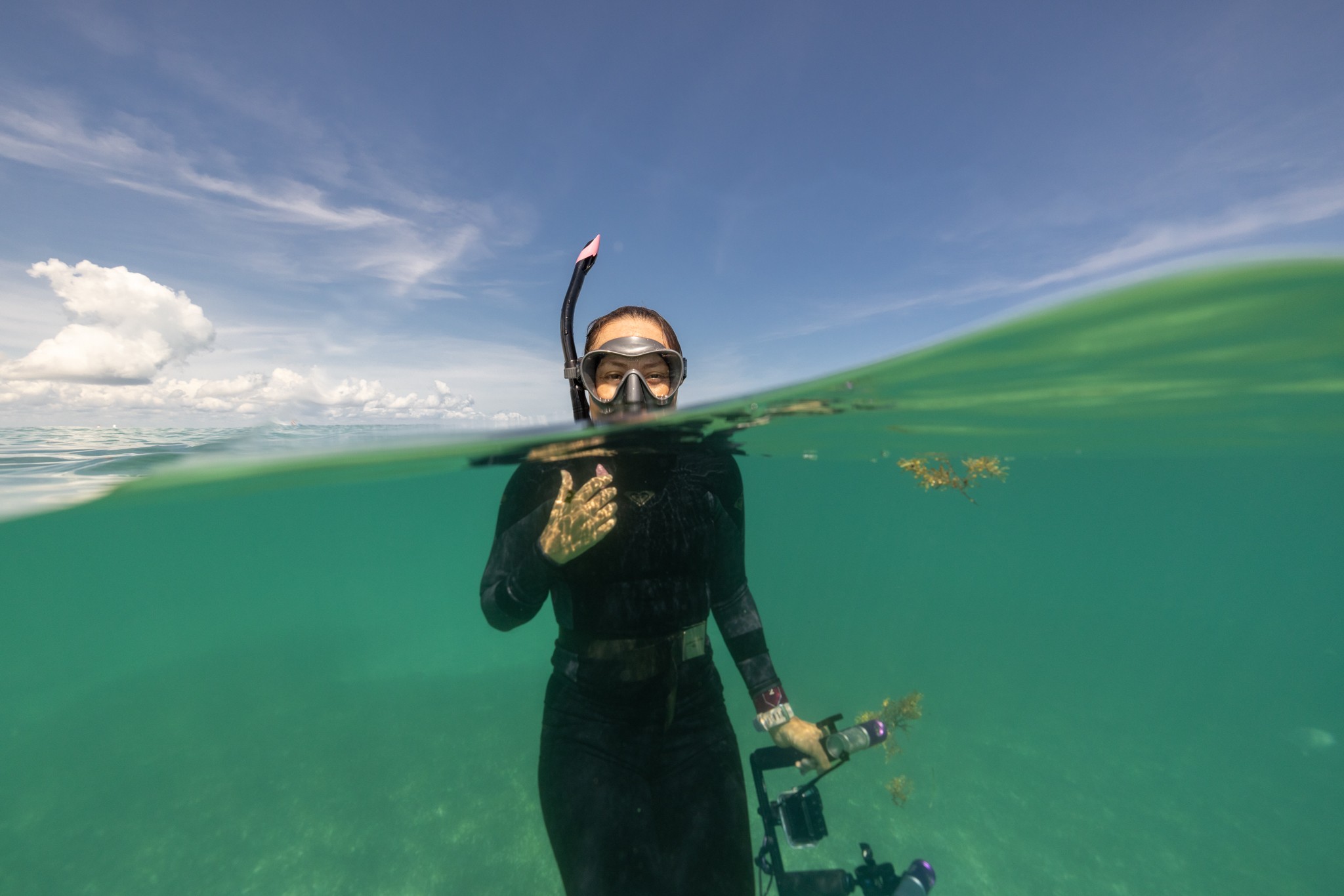 Person in wetsuit with snorkel and mask holding a camera underwater, waving above water, with blue sky and clouds.