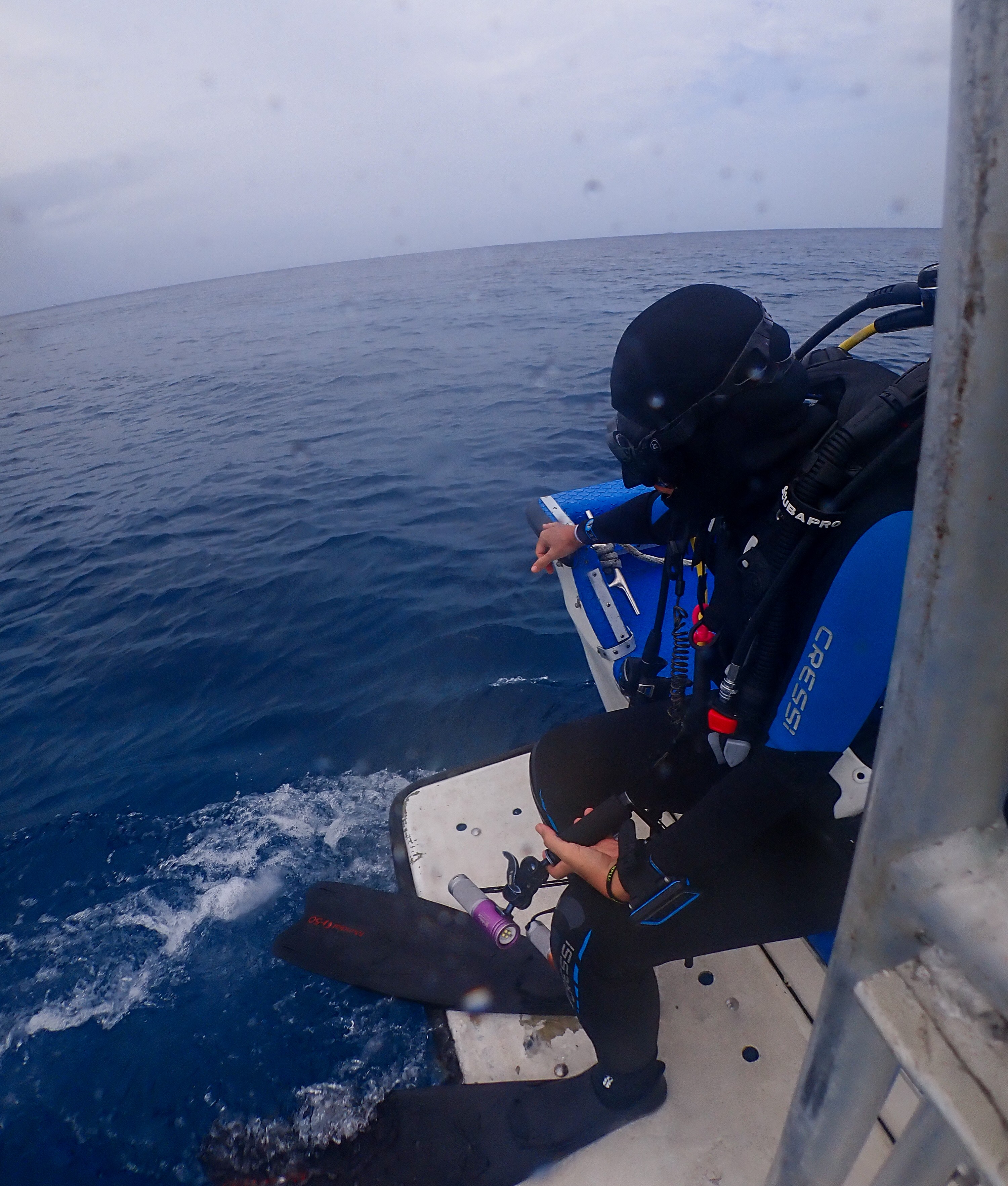 Person in wetsuit and helmet on boat, preparing to jump into the ocean, with water splashing below.