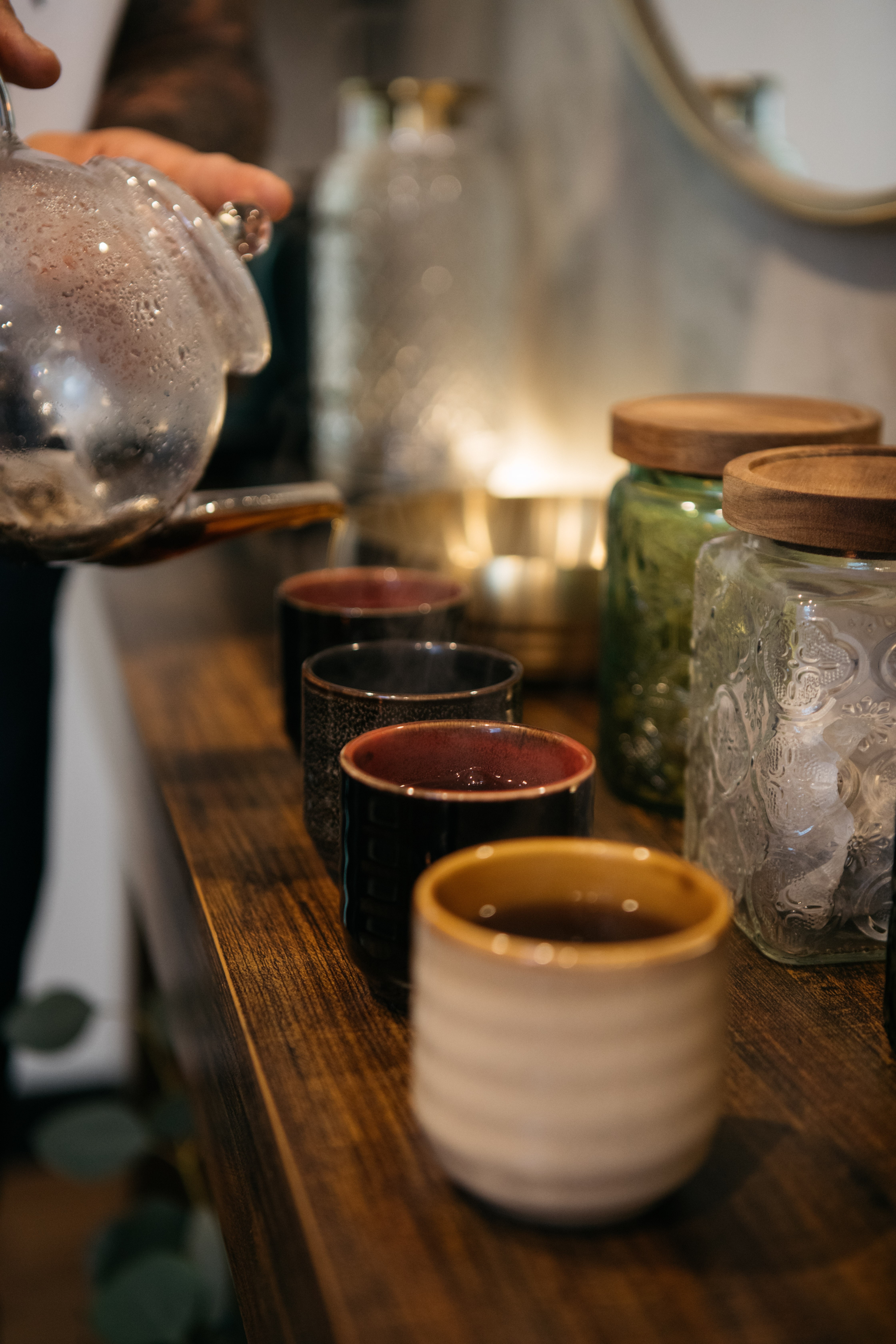 Row of cups and jars on a wooden table with a blurred background, including a glass teapot and containers.
