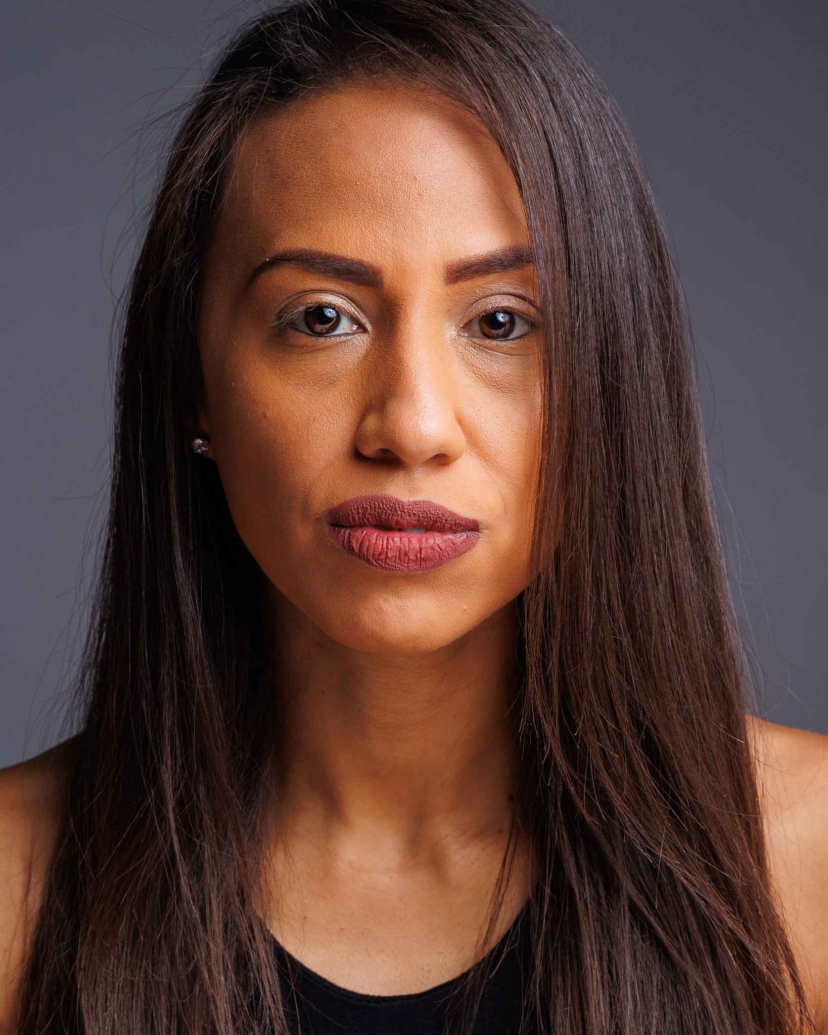 Portrait of a woman with long dark hair, neutral expression, wearing a black top, against a gray background.