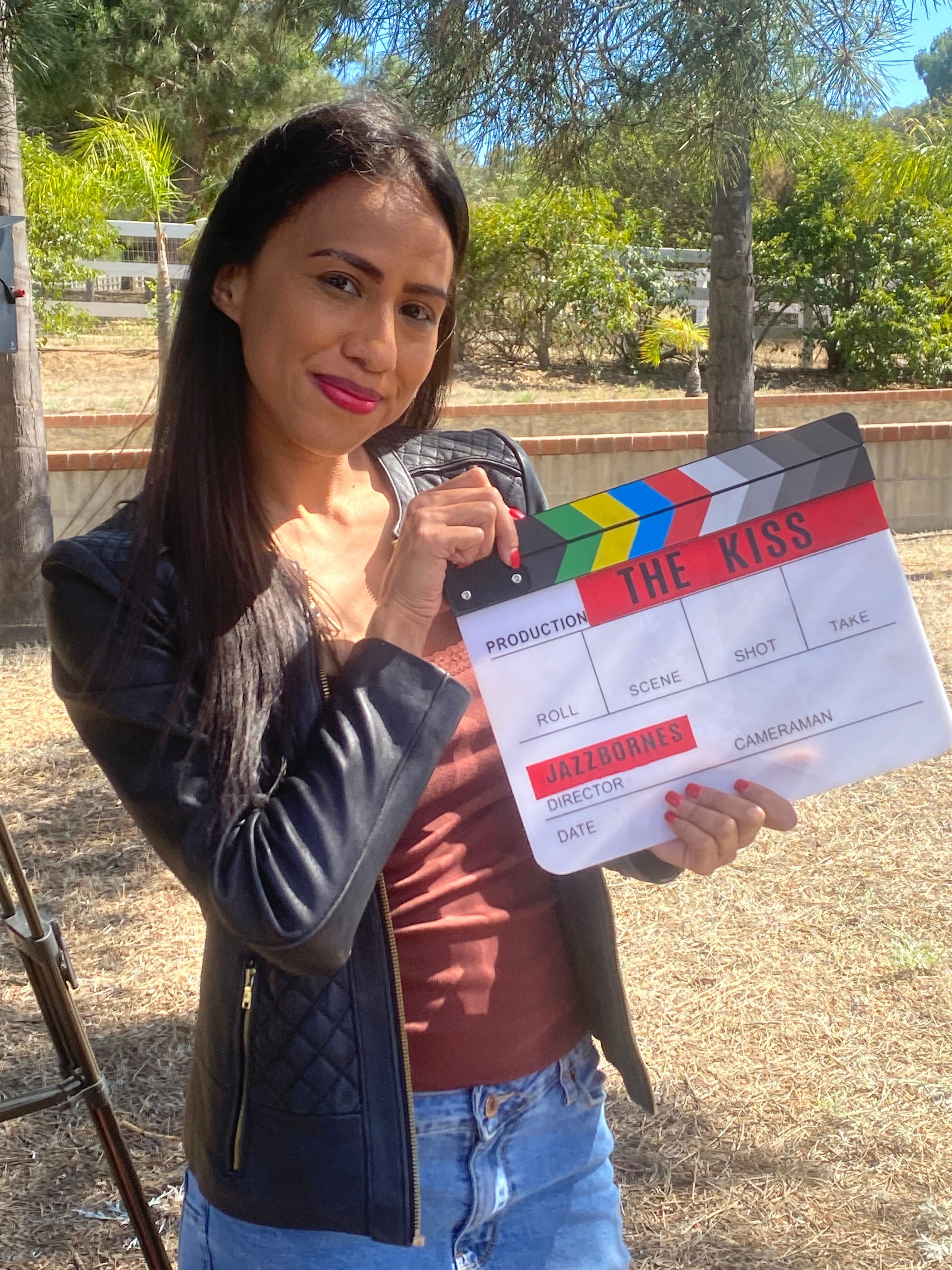 Woman holding a clapperboard and a microphone outdoors, smiling, with trees and a pathway in the background.