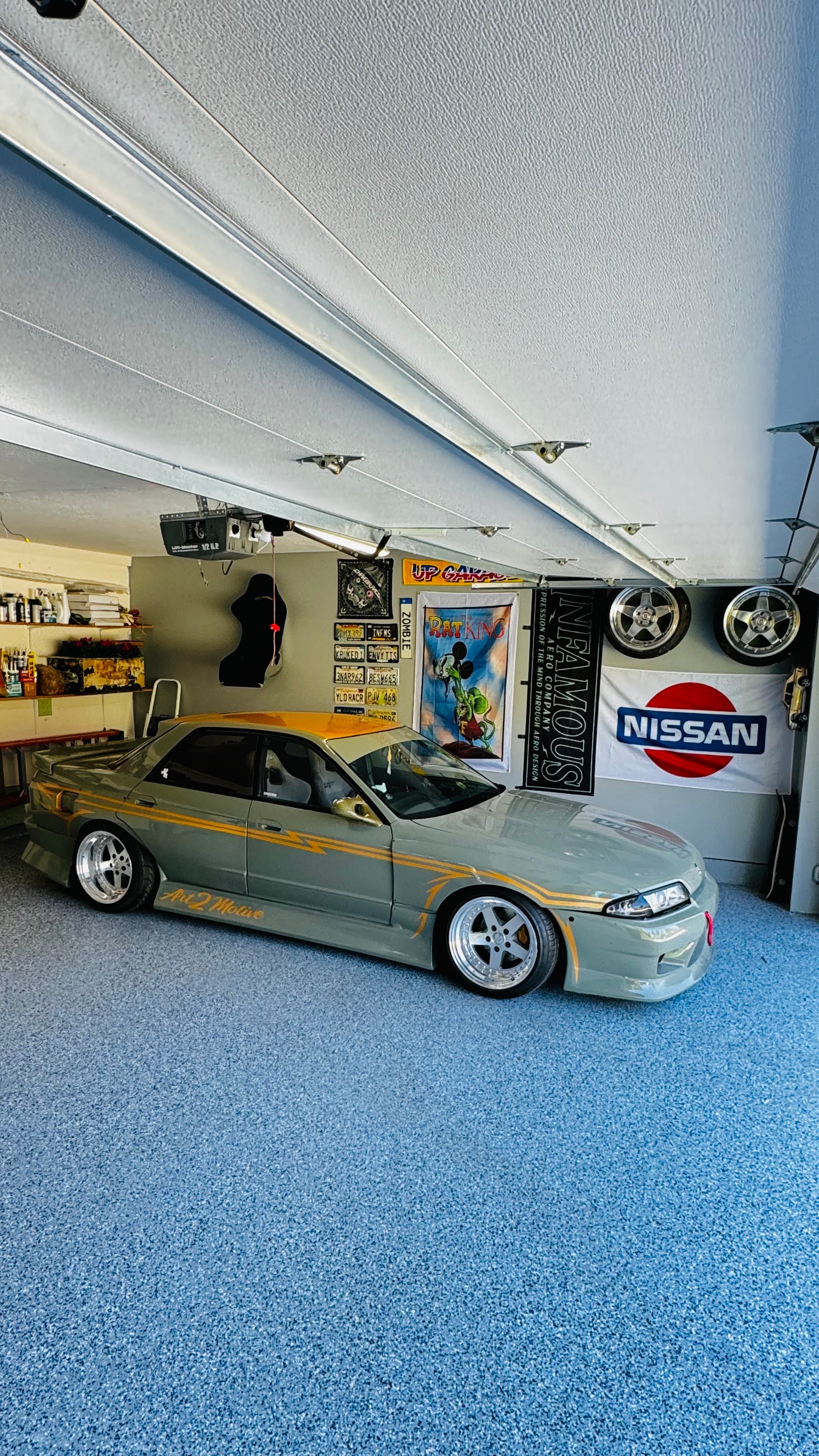Model car displayed indoors with Nissan sign and posters, blue carpeted floor, and ceiling with lighting fixtures.