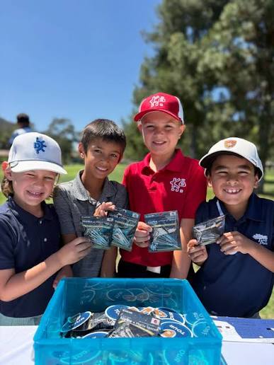 Four children smiling outdoors, holding packaged items, with a blue container in front, trees and sky in background.