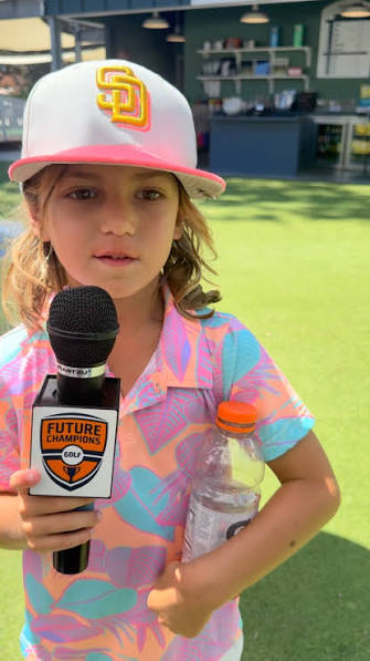 Young girl holding a microphone with a logo, wearing a baseball cap and colorful shirt, outdoors.