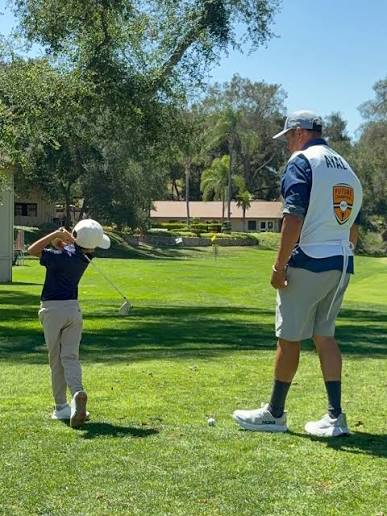 Child and adult on a golf course, child holding a golf club, adult watching, trees and blue sky in background.