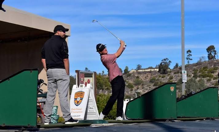 Person swinging golf club at driving range with instructor nearby, green barriers, and a scenic hillside background.