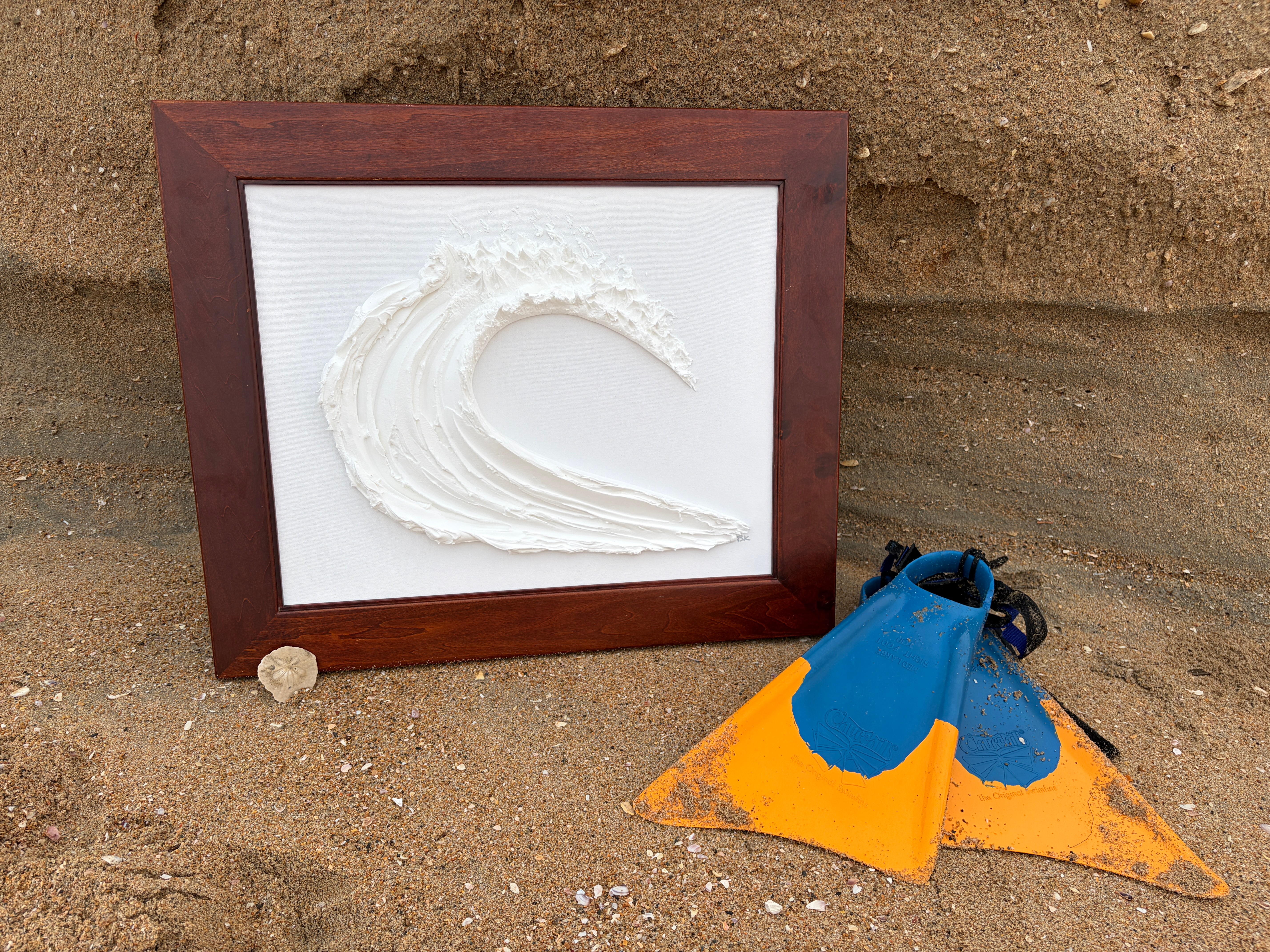 Framed artwork with a white textured wave on a white background, placed on sandy ground with a blue and orange object nearby.