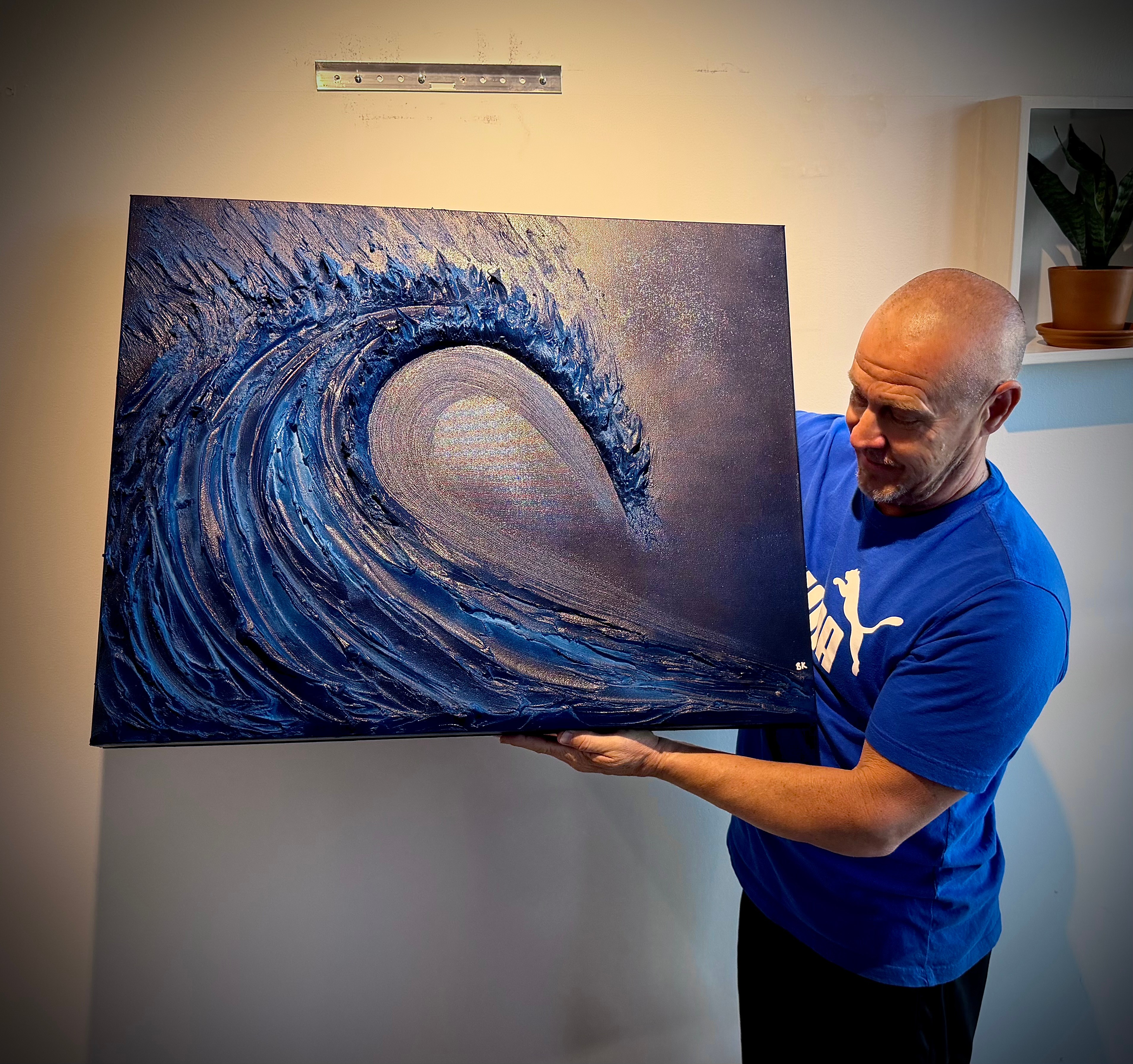 Man holding a large painting of a wave, standing indoors near a wall with a potted plant on a shelf.