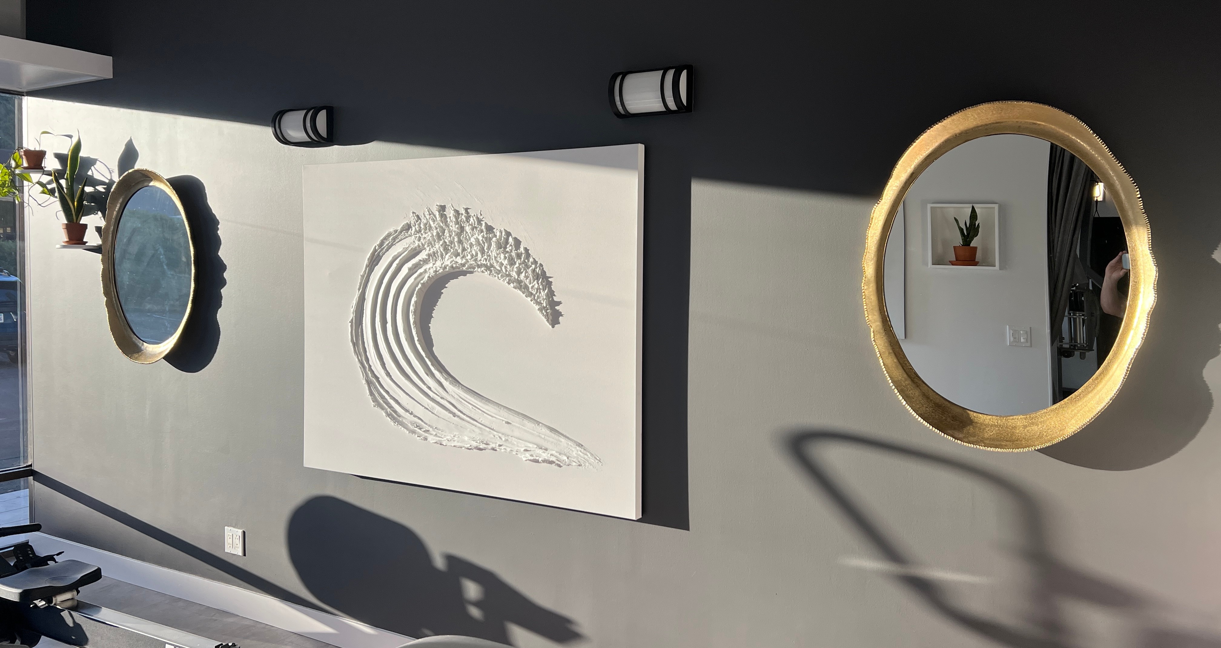 Gallery wall with three circular mirrors and a wave artwork on a gray wall, sunlight casting shadows.