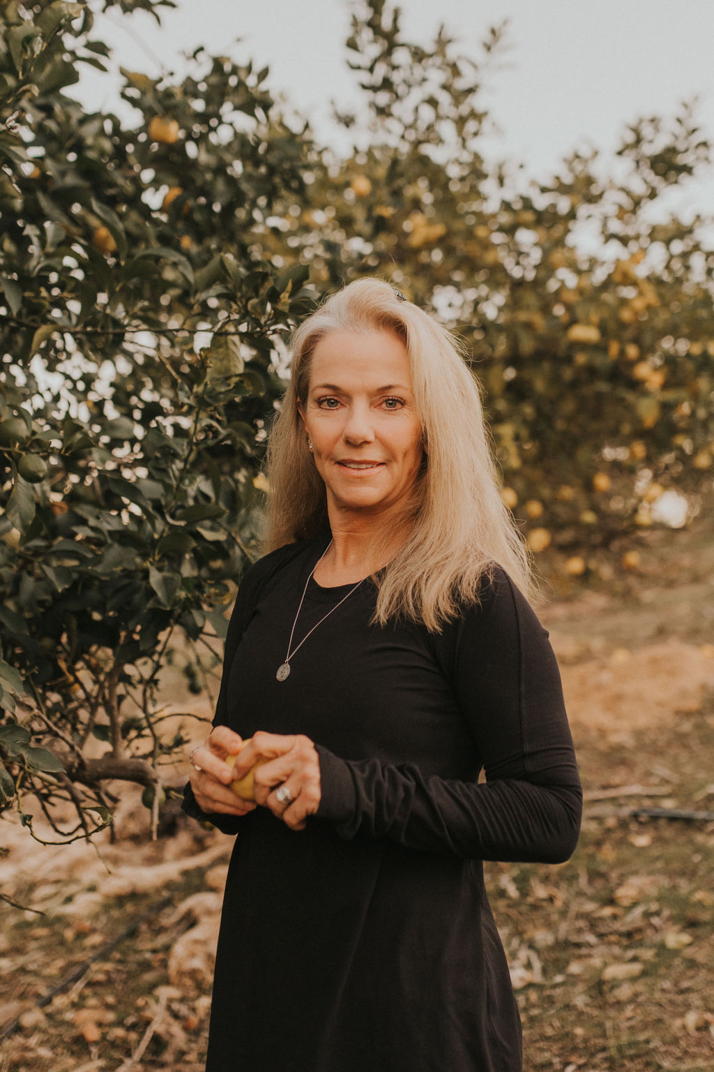 Woman with long blonde hair standing outdoors near trees, smiling, wearing a black long-sleeve top, holding her hands together.