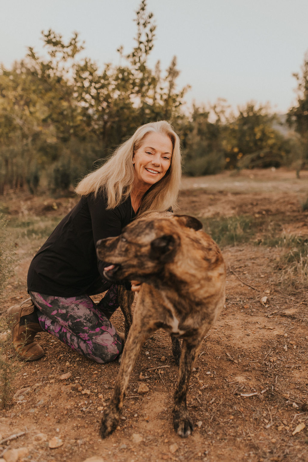 Woman smiling and hugging a large dog outdoors on dirt ground with trees in background.