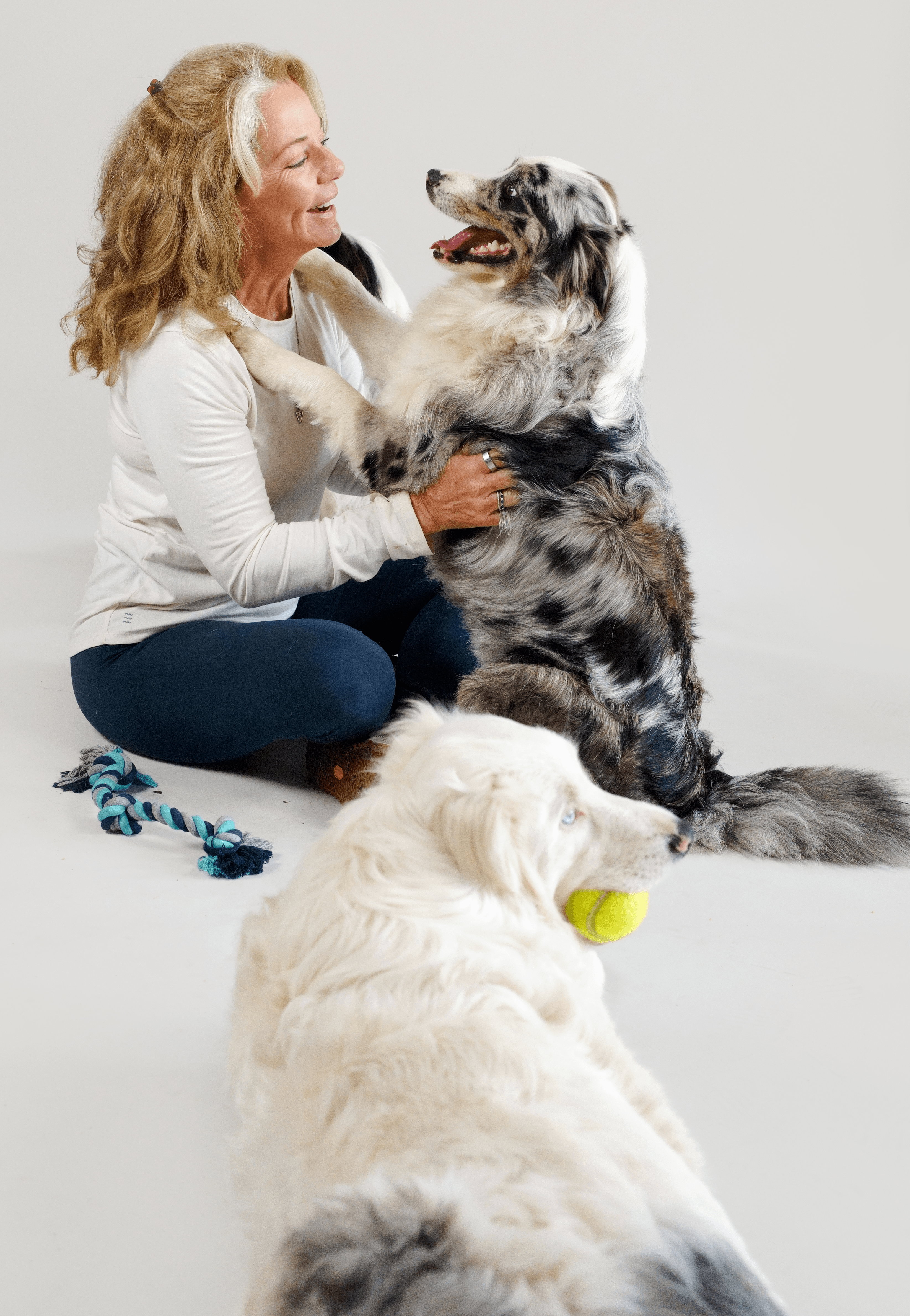 Woman with curly hair playing with two dogs, one sitting and one lying down with a yellow ball, against a plain background.