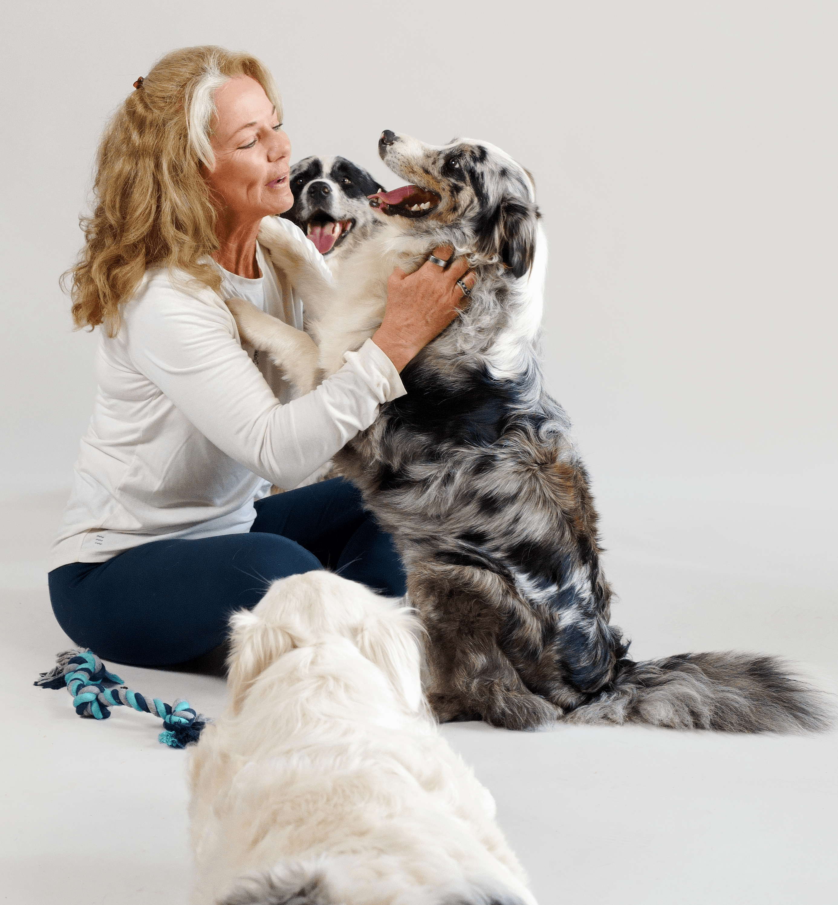 Woman with curly hair playing with two dogs, one sitting and one lying down, against a plain background.