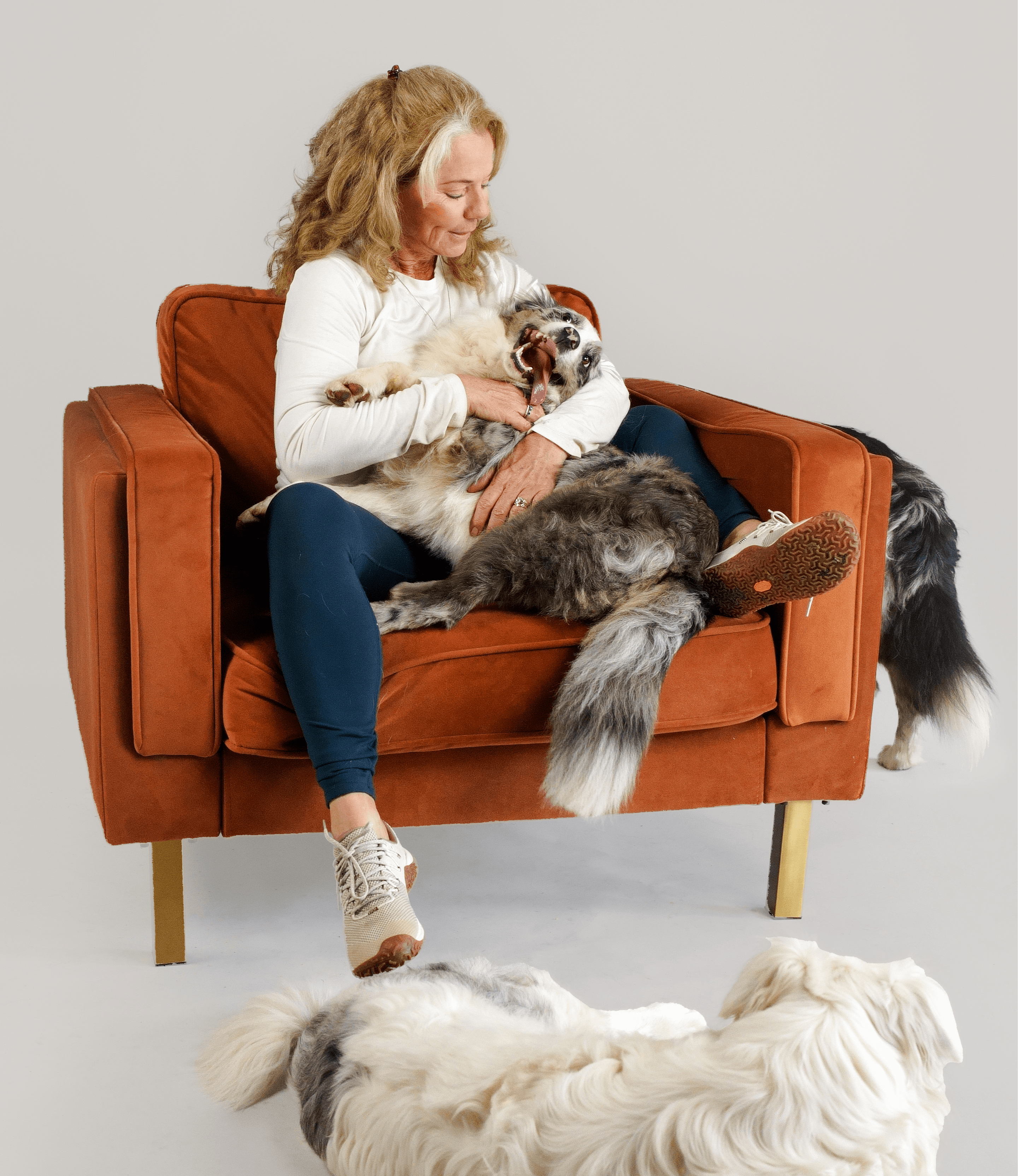 Woman sitting on a brown sofa holding a large dog, with another dog lying on the floor nearby, against a plain background.
