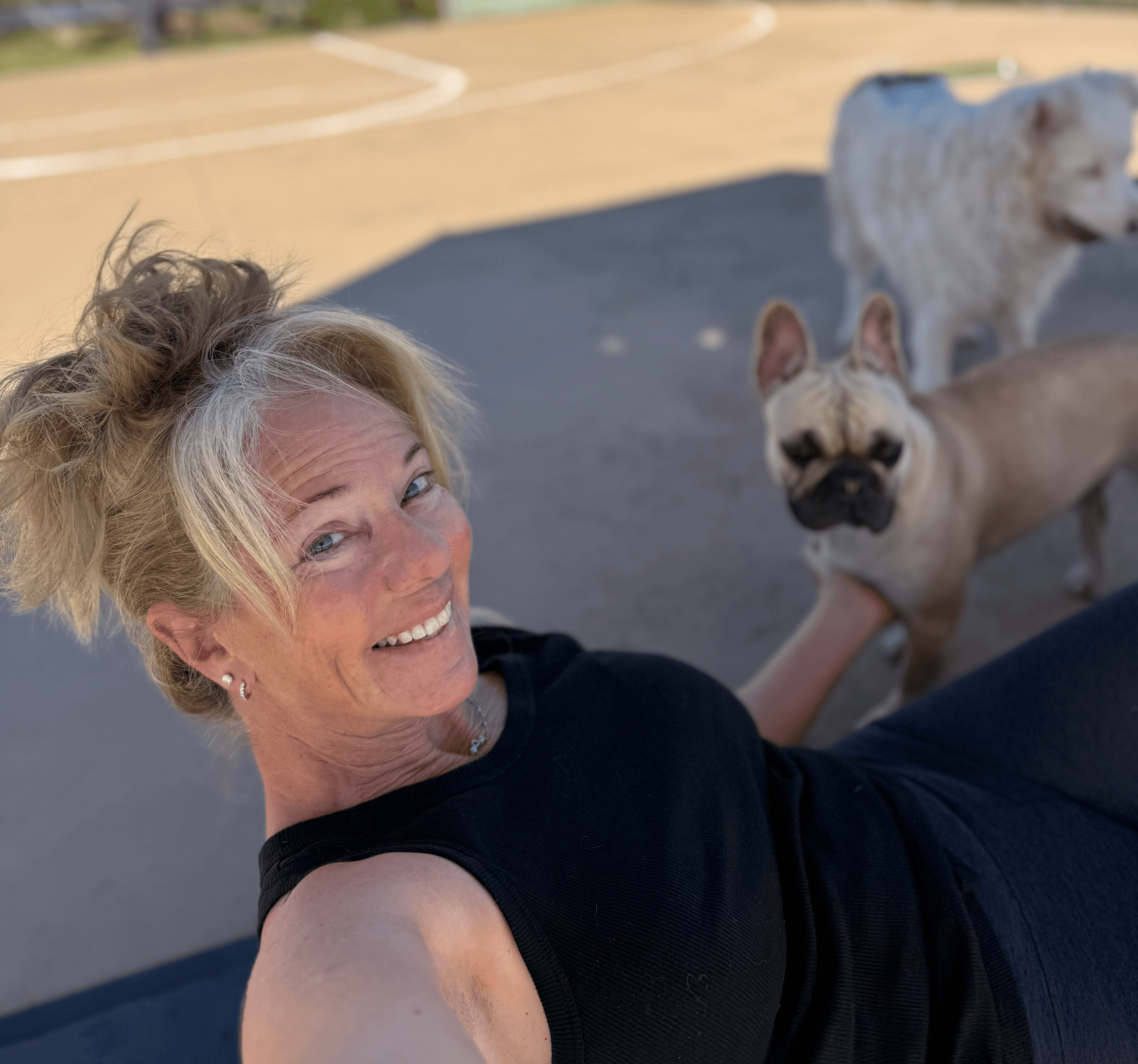 Woman with blonde hair smiling outdoors with three dogs, one white and fluffy, one small and tan, and one medium-sized, on a paved surface.