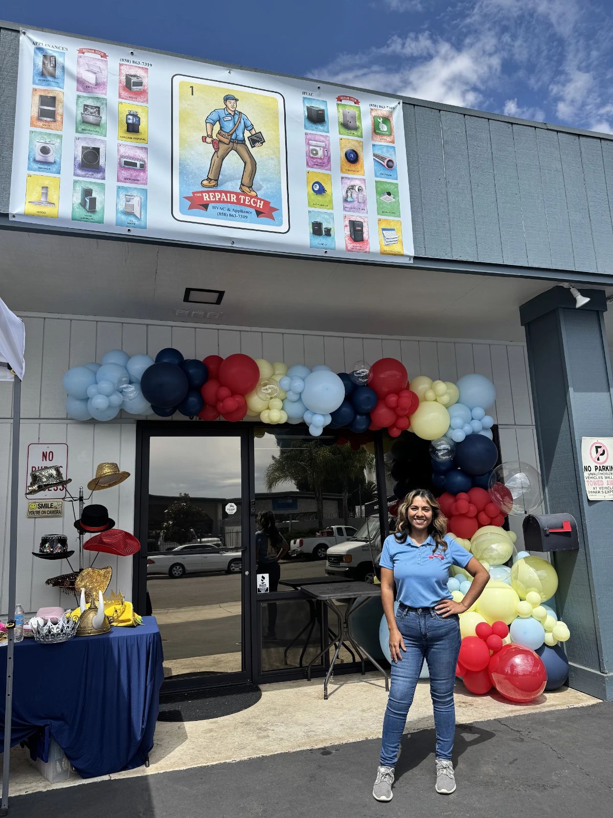 Woman standing outside store entrance decorated with balloons, with a table of party favors and a poster above.