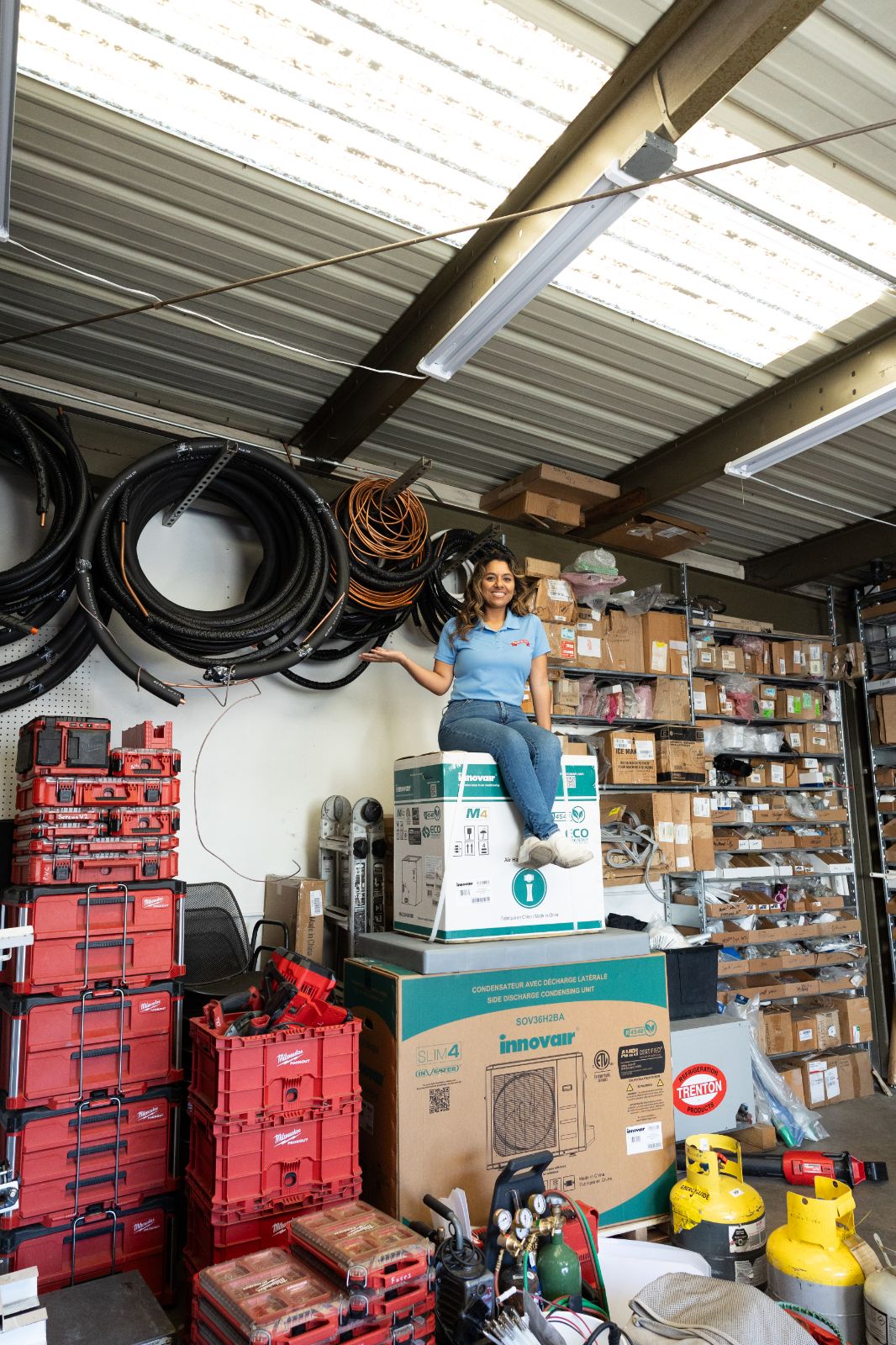 Woman standing on a box in a warehouse with shelves, boxes, and tools around her.