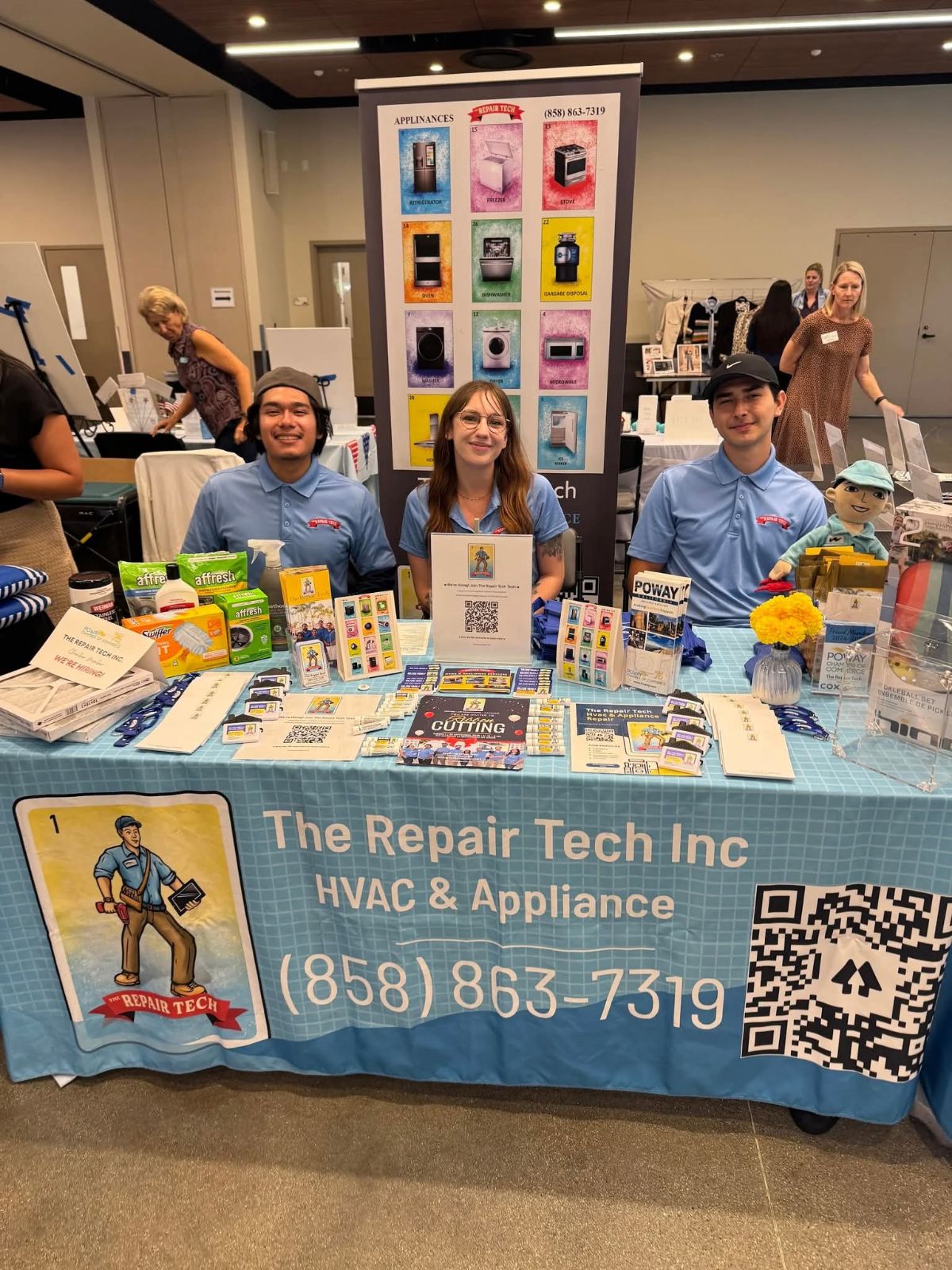Three people sit behind a table with HVAC and appliance repair products, a banner displays contact information, and a display board is behind them.