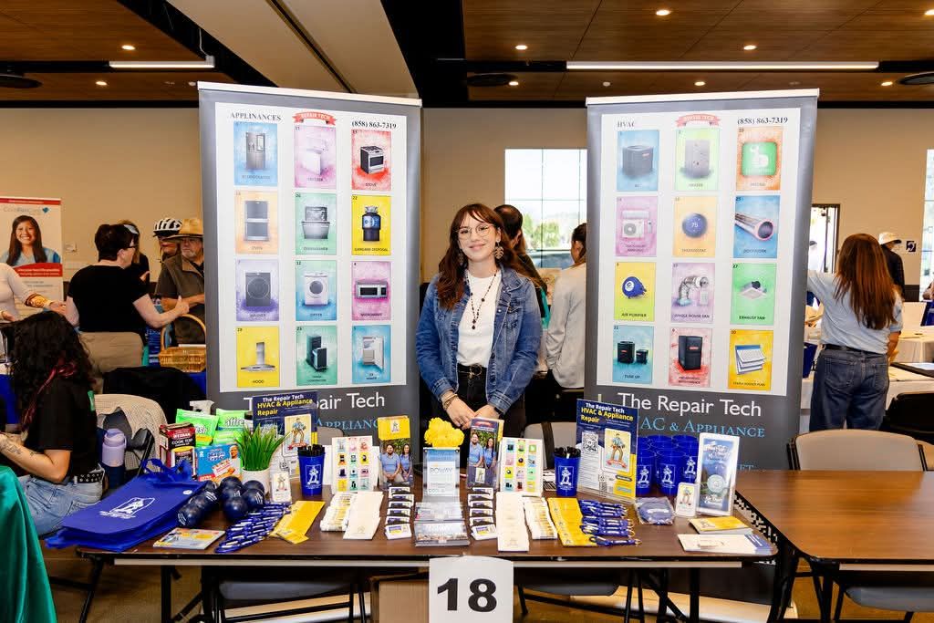 Woman standing behind a table with repair tools and products, banners in the background, at an indoor event or exhibition.