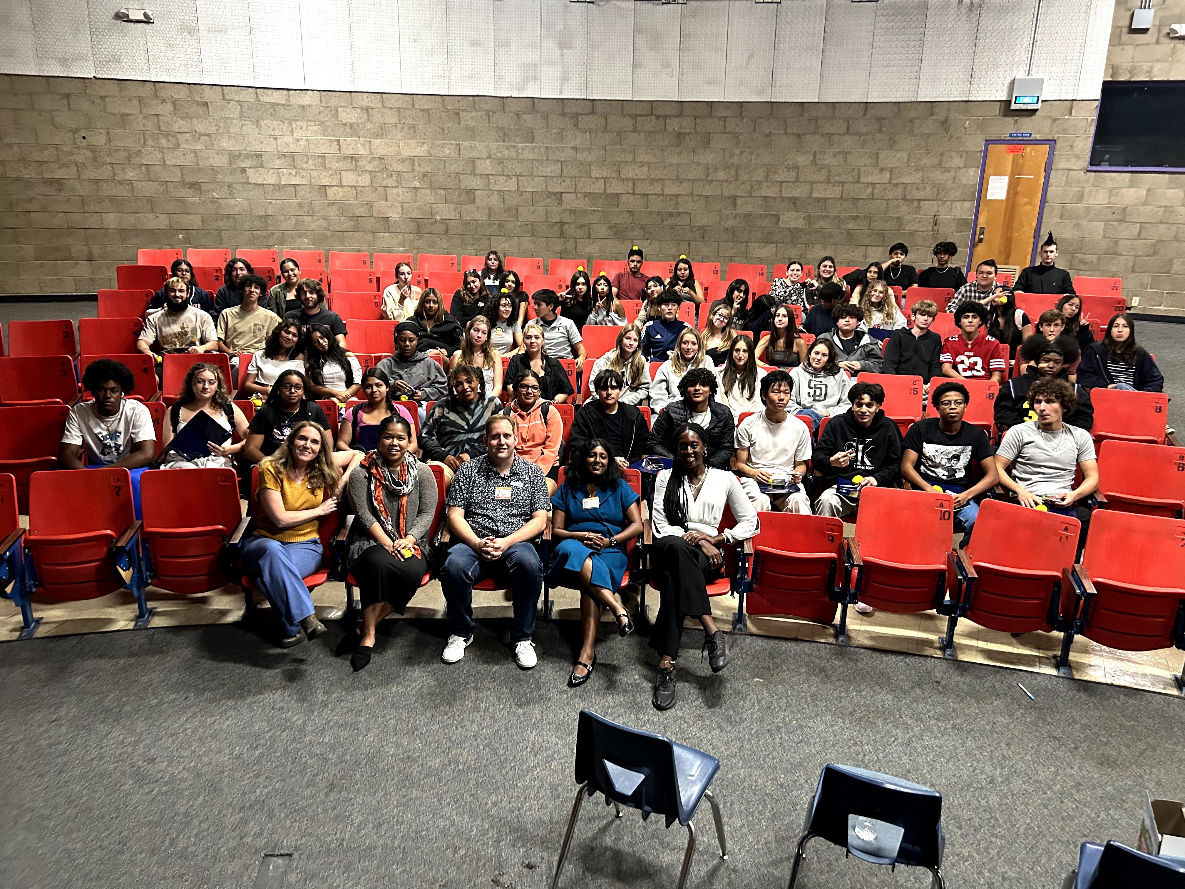Group of children and adults seated in red chairs in an auditorium, facing forward, with a beige wall background.