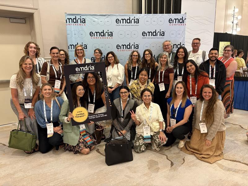 Group of people posing in front of a backdrop with 'emdria' logo, some holding signs, indoors, smiling, diverse group.