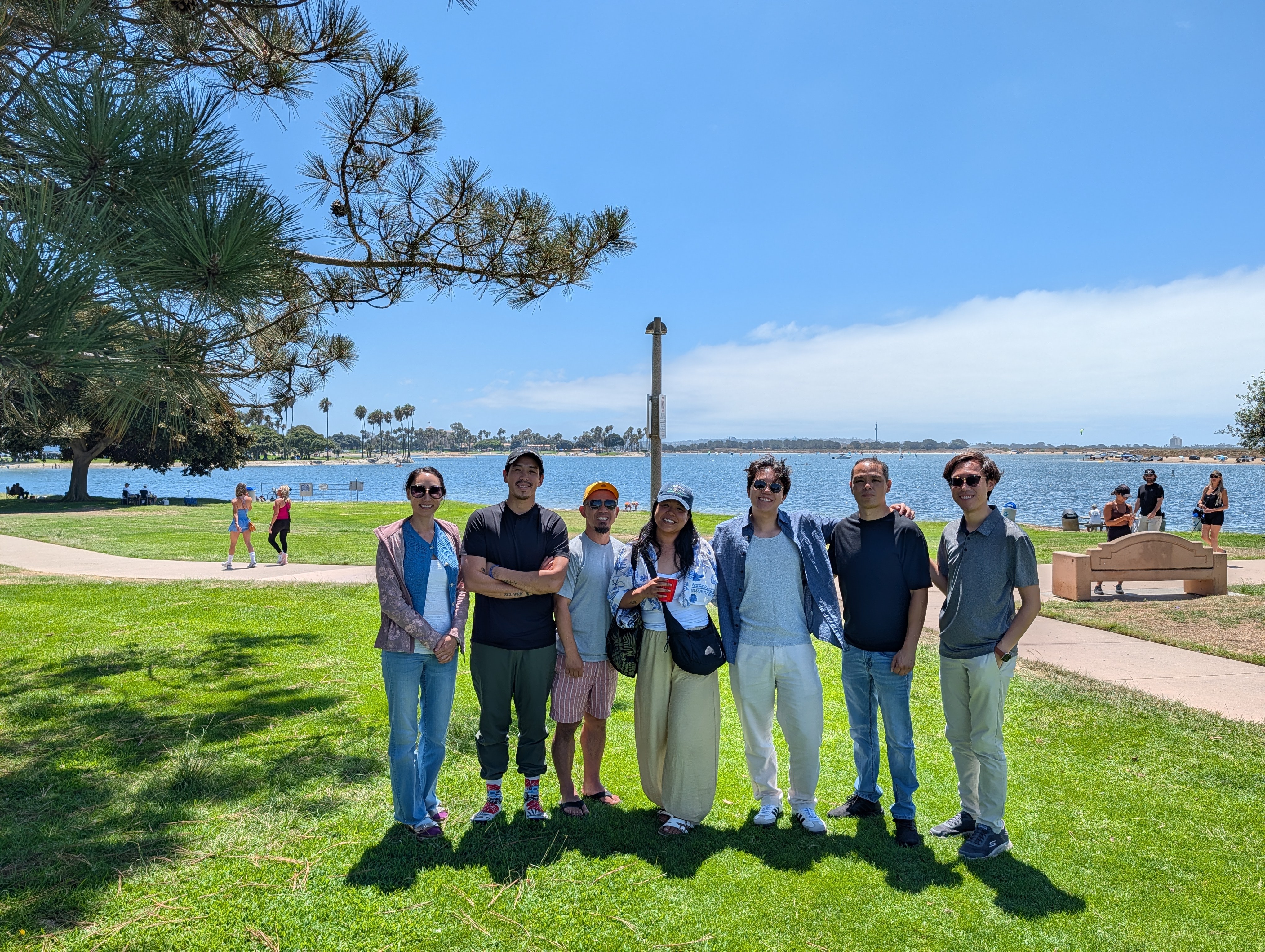Group of seven people standing on grass near water with trees and a walkway in the background.