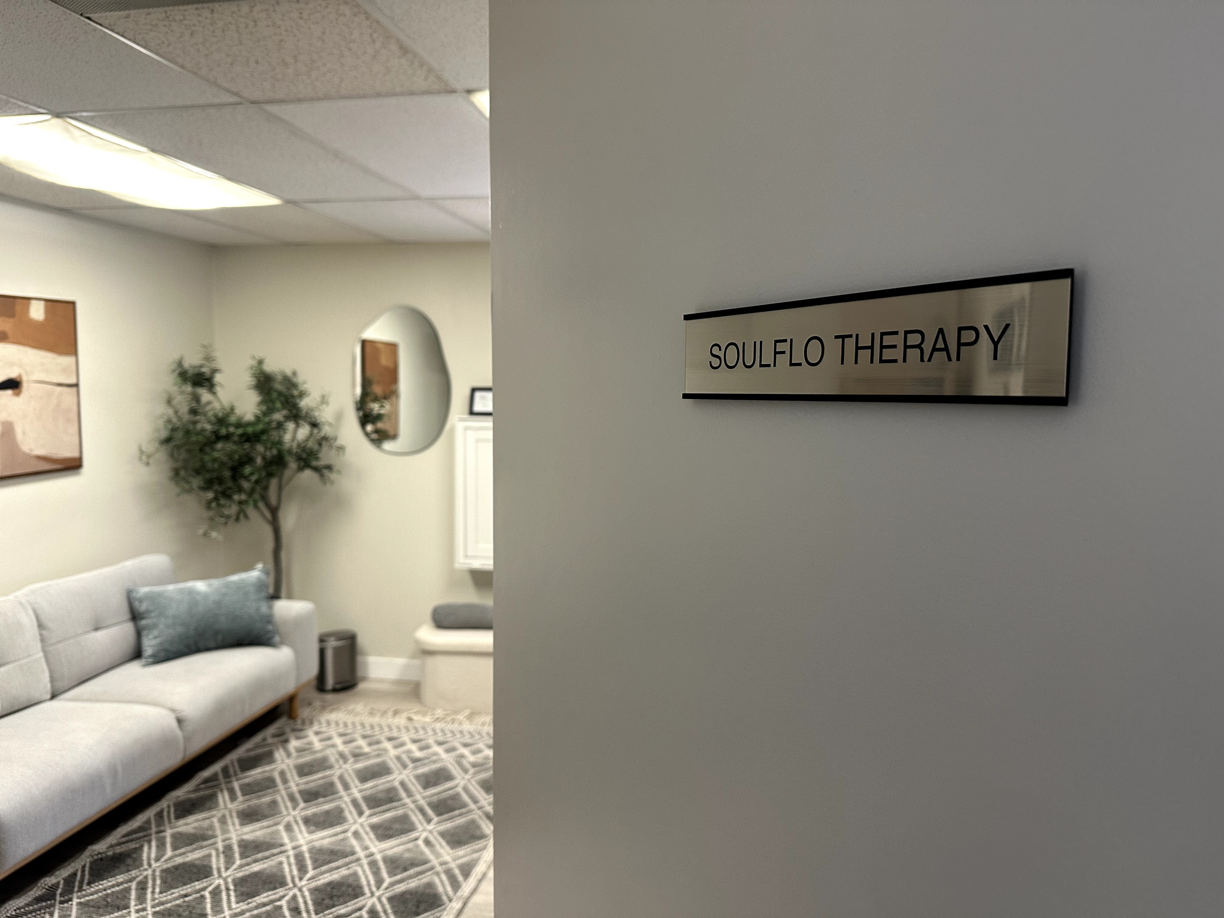 Waiting room with a white sofa, a small tree, and a sign reading 'SOULFLO THERAPY' on the wall.