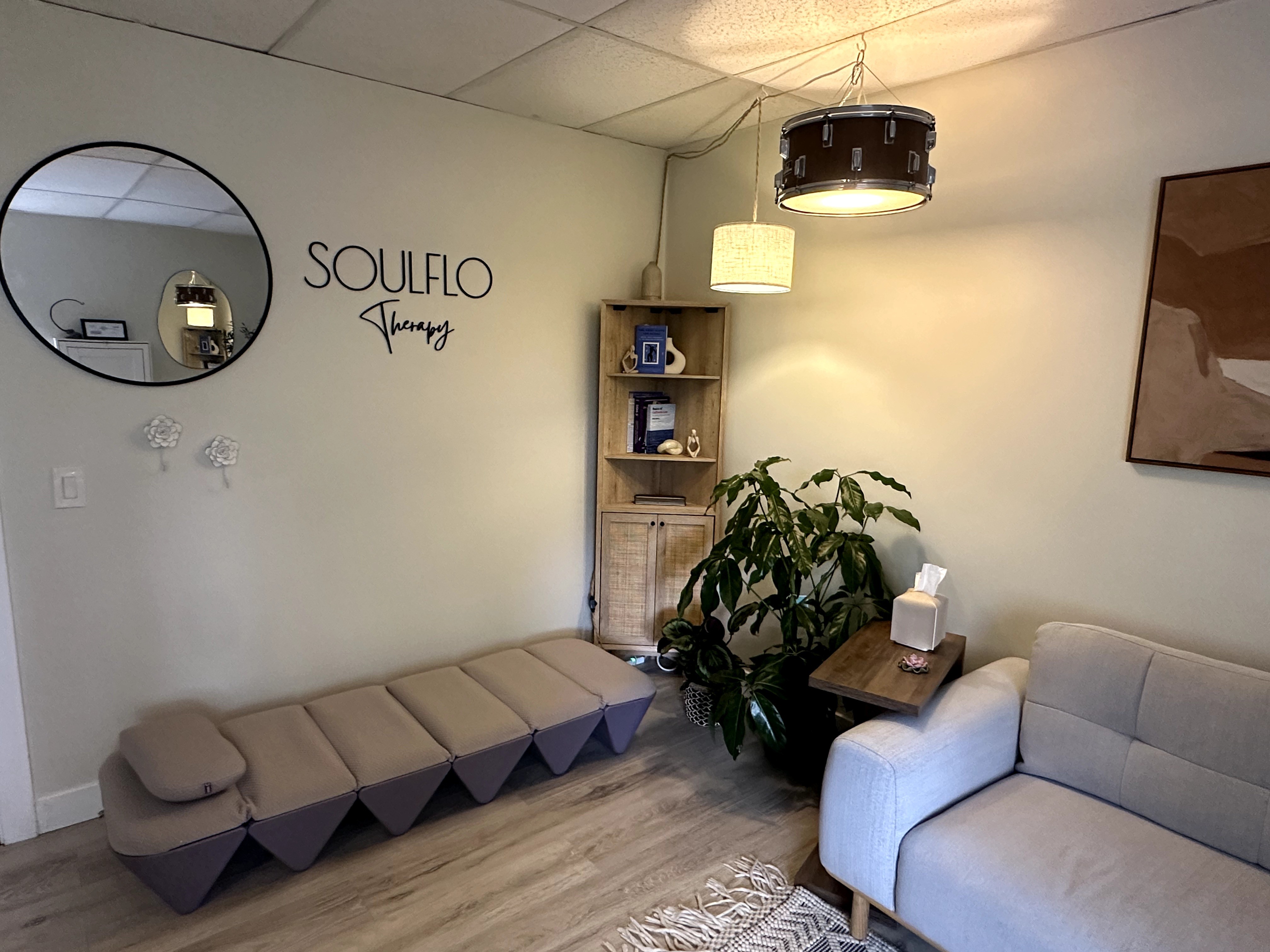 Living room with a beige sofa, a wooden shelf, a large plant, and a wall mirror with the words 'SOULFLO' and 'Viny' written nearby.