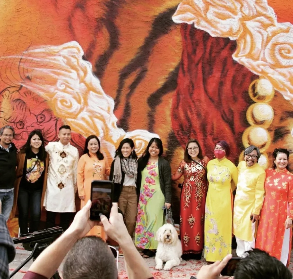 Group of women in traditional dresses standing in front of a large tiger mural, some taking photos.