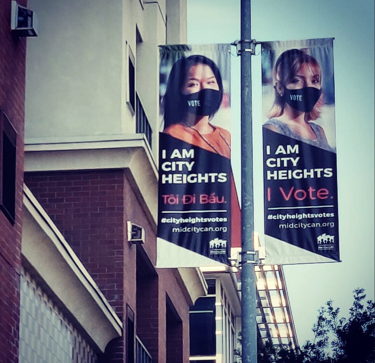 Two vertical banners on a street pole feature women wearing face masks with text about voting and city heights.