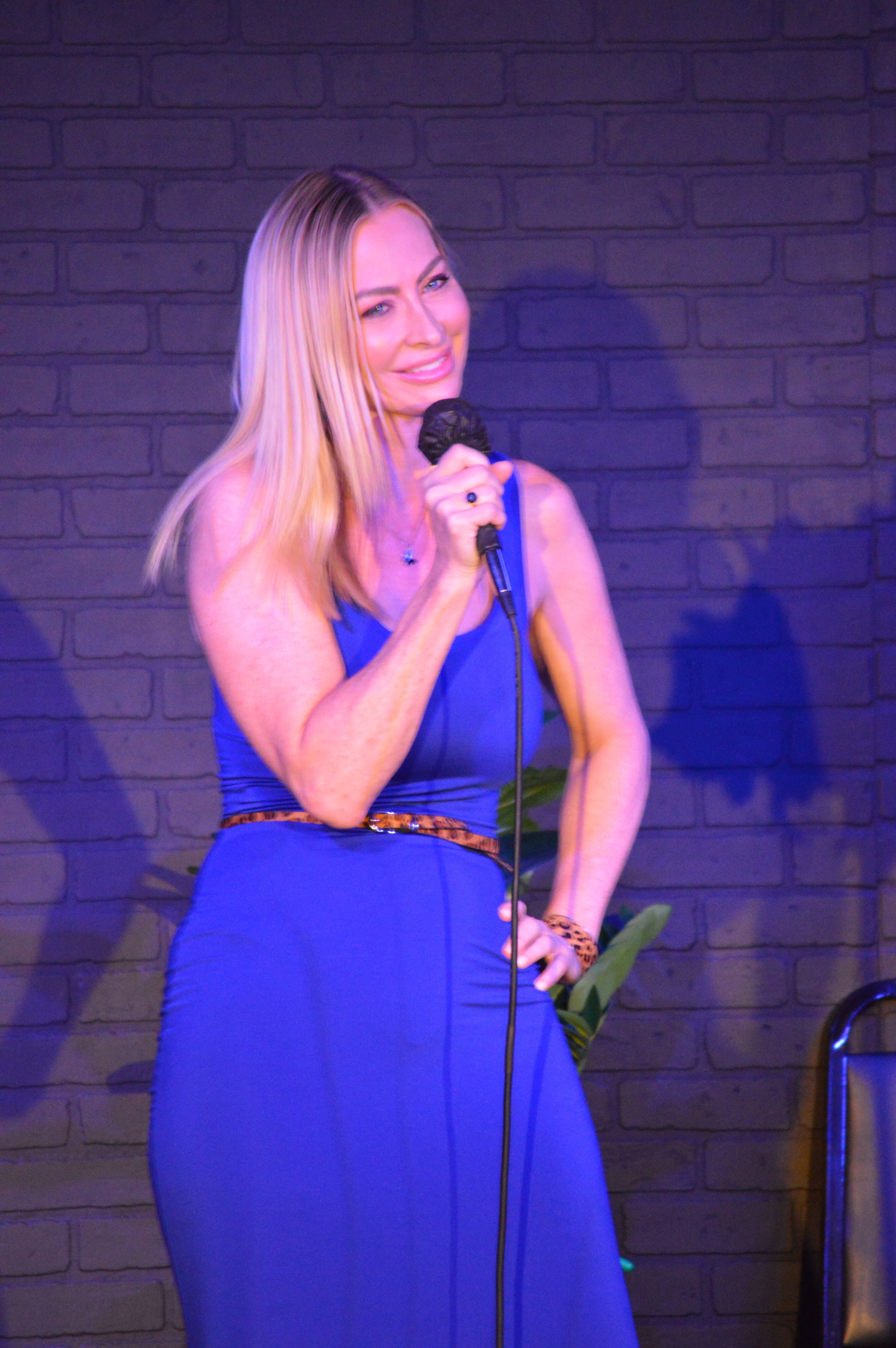 Woman with long blonde hair in a blue dress holding a microphone and a rose, singing on stage against a brick wall.