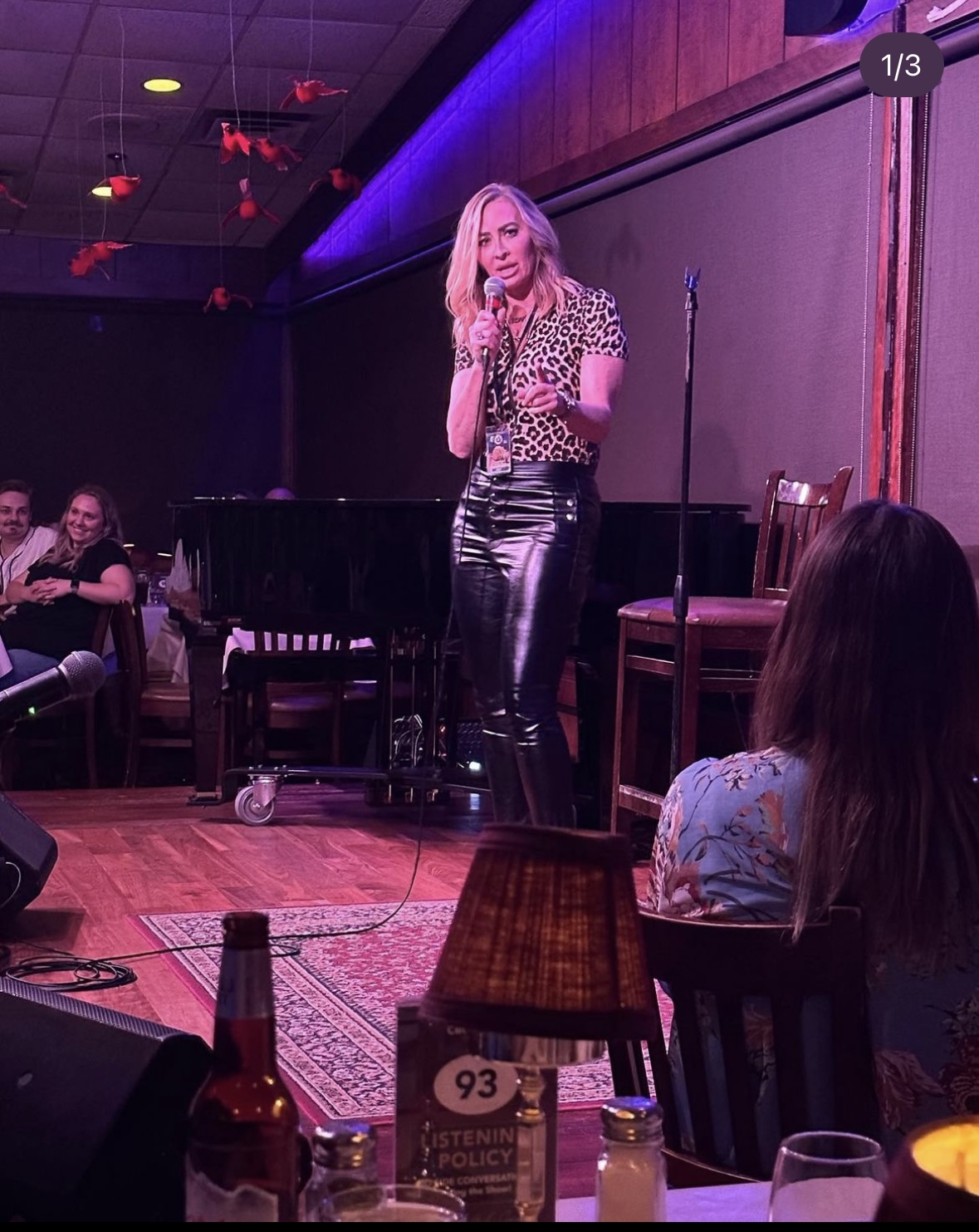 Woman performing on stage with microphone, audience seated, wooden floor, dim lighting, and a patterned rug.