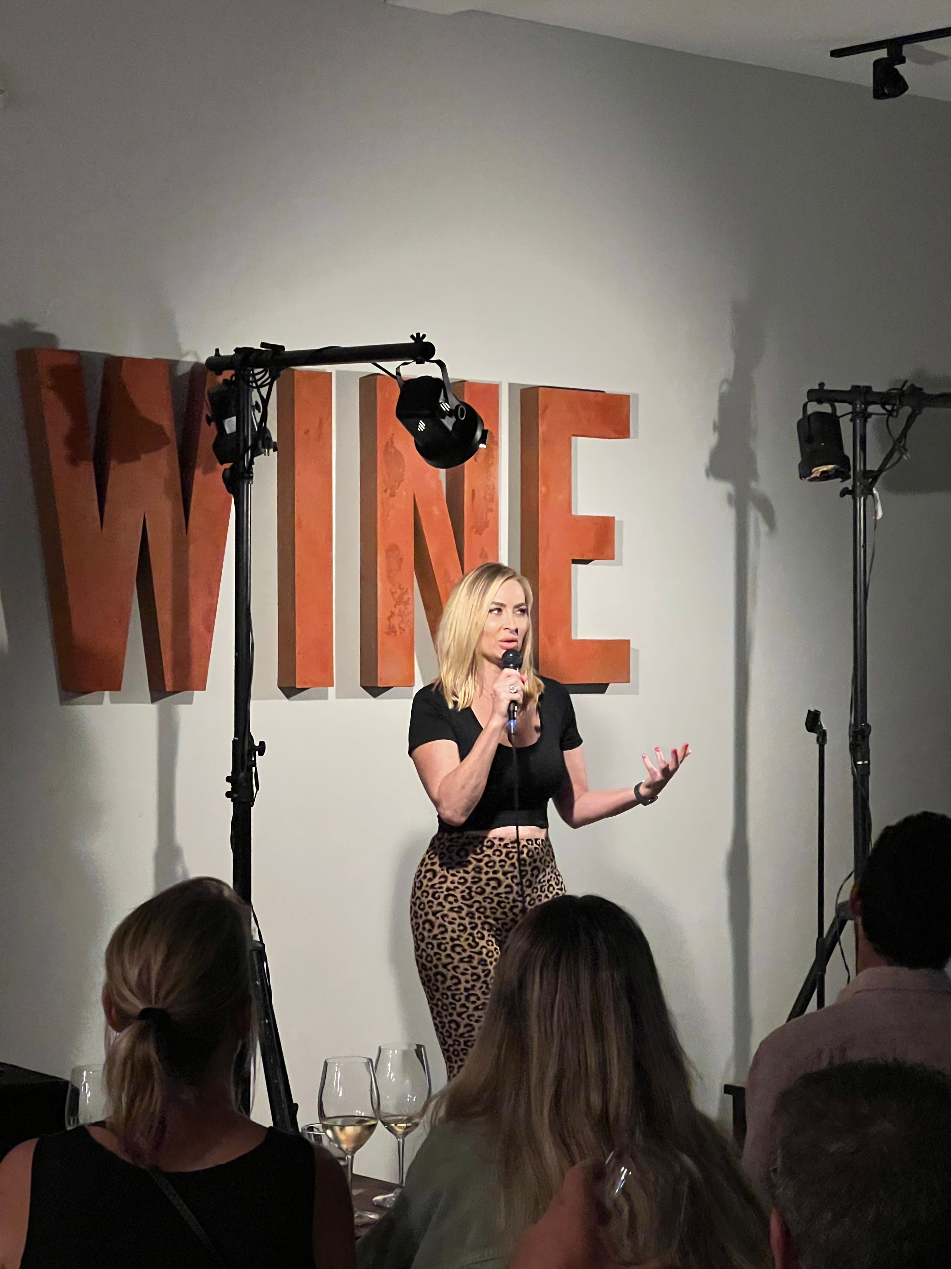 Woman performing stand-up comedy on stage with a microphone, audience watching, and large orange letters in the background.