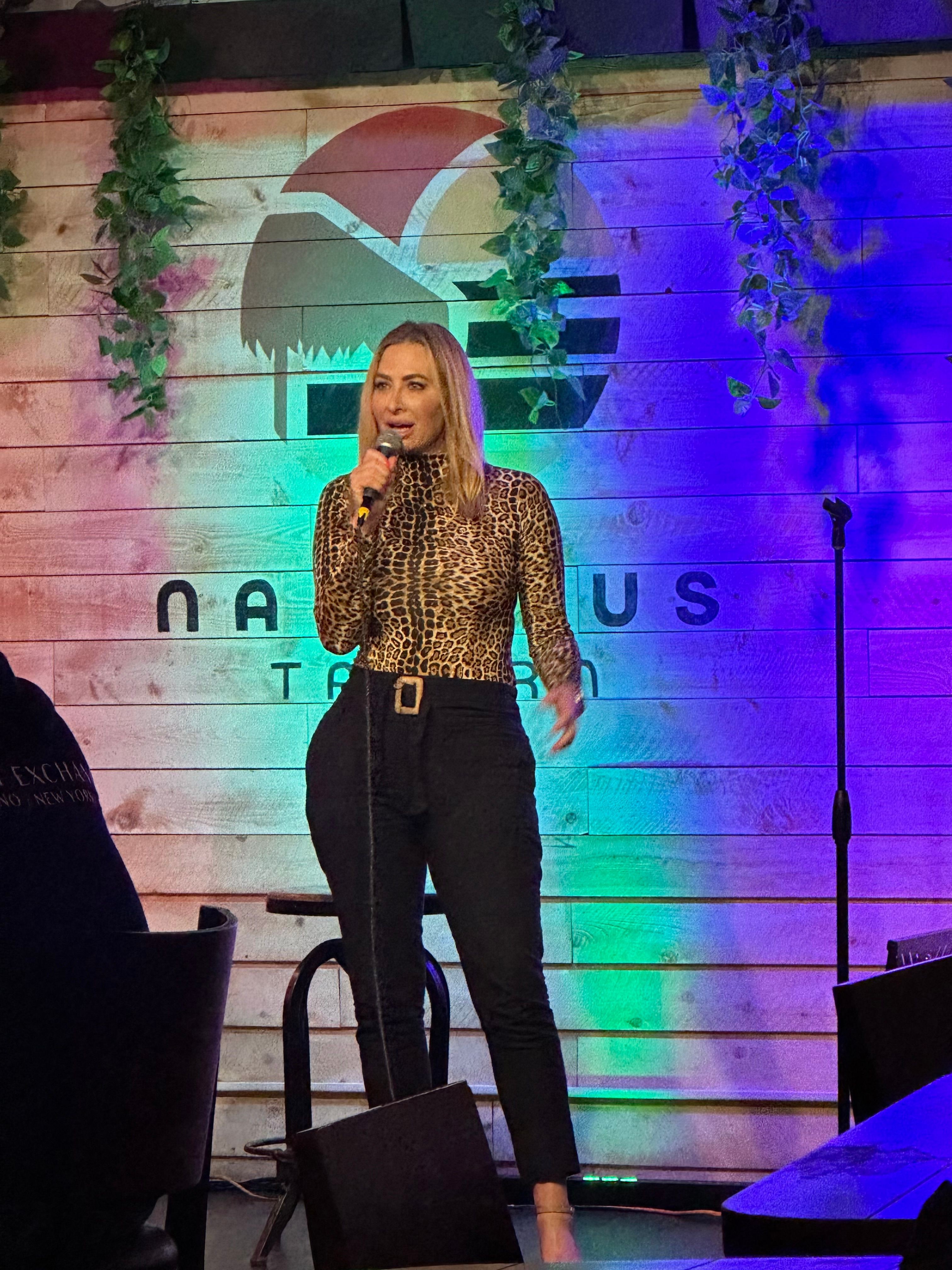 Woman standing on stage with colorful background, wearing a leopard print top and black pants, gesturing with her right hand.
