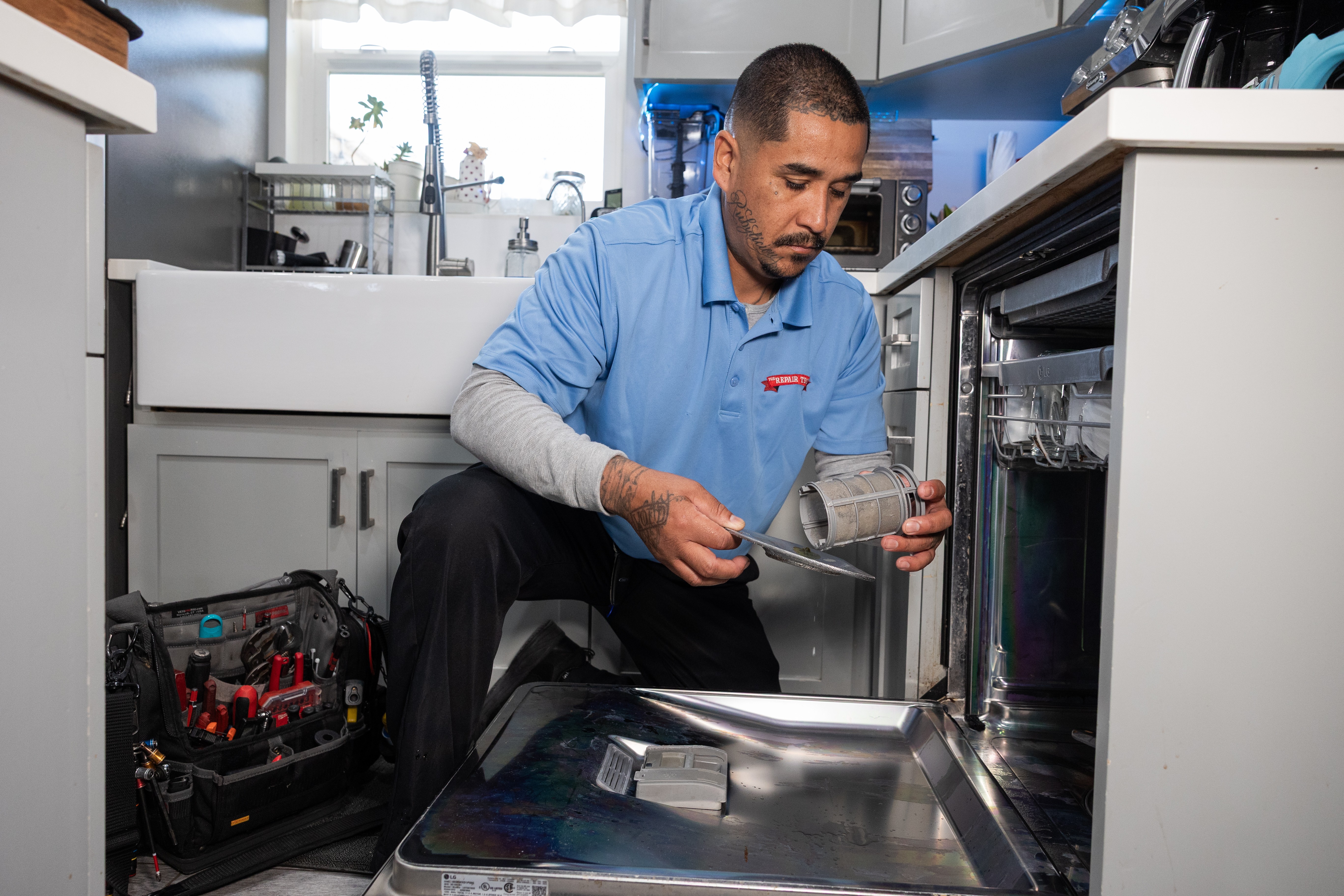 Man in blue shirt kneeling in kitchen, holding a glass container near dishwasher, with kitchen cabinets and appliances around.