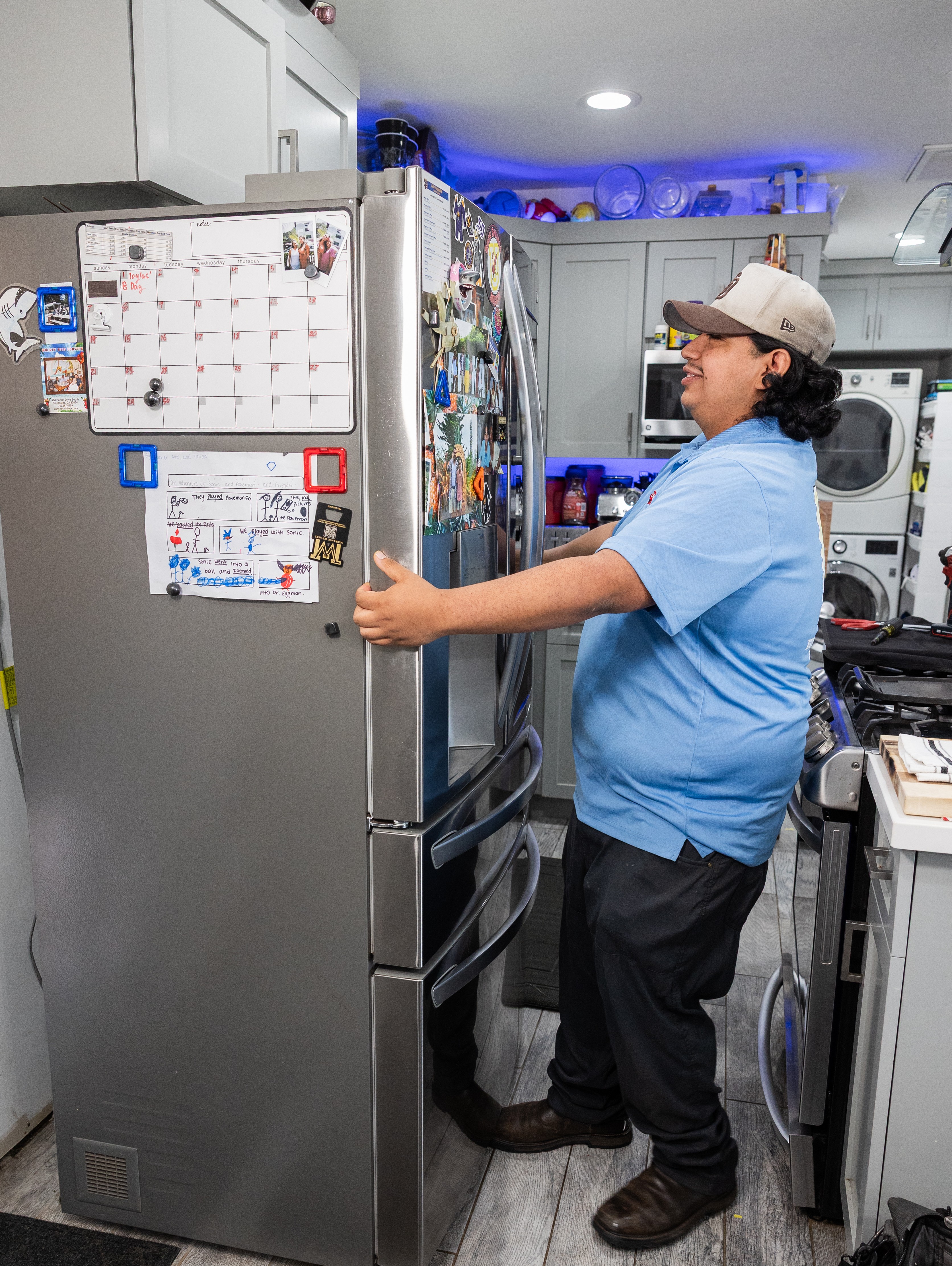 Person in blue shirt and cap standing in kitchen, reaching for a refrigerator door, with appliances and shelves around.
