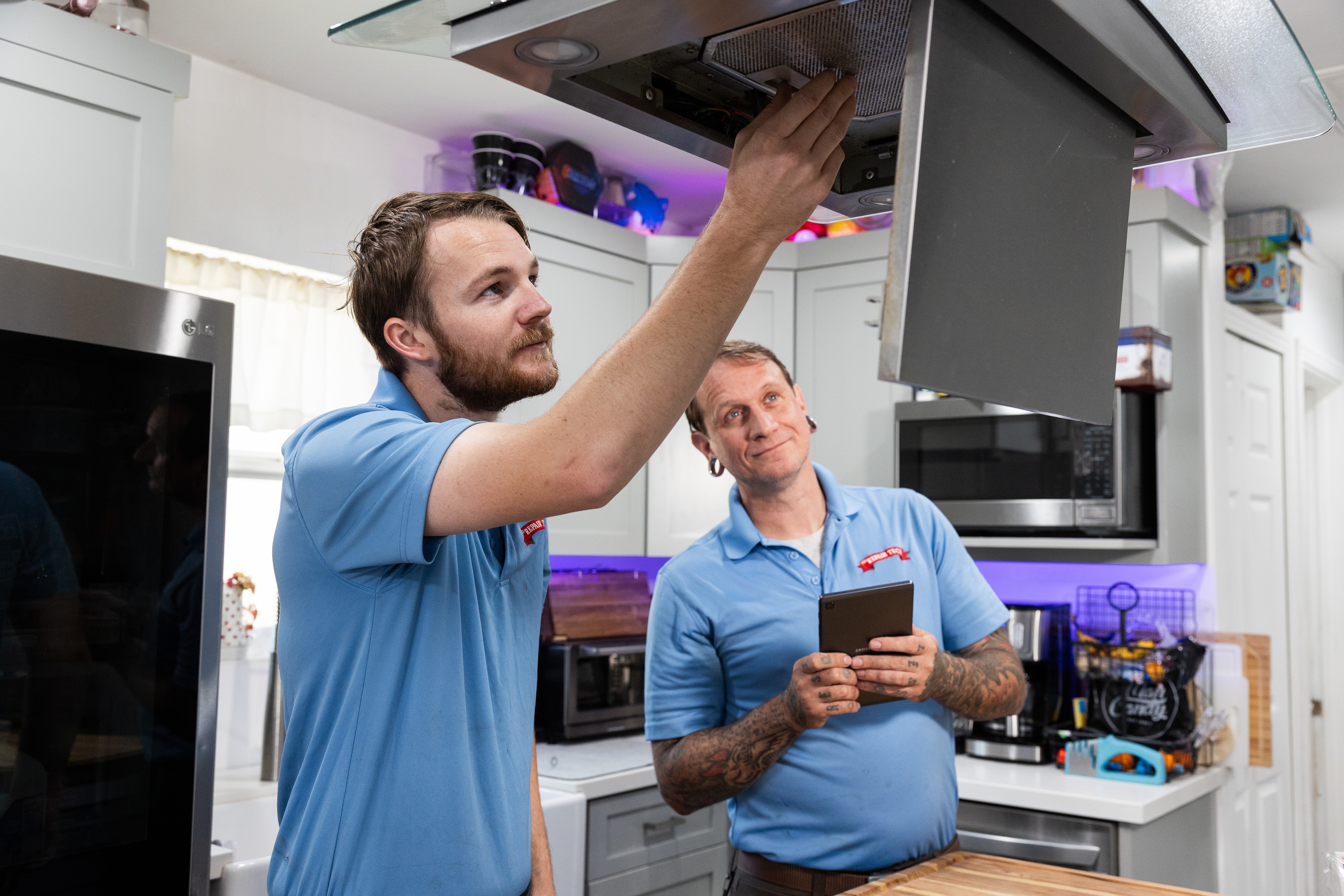 Two men in blue shirts working in a modern kitchen, one reaching into an overhead cabinet, the other holding a phone.