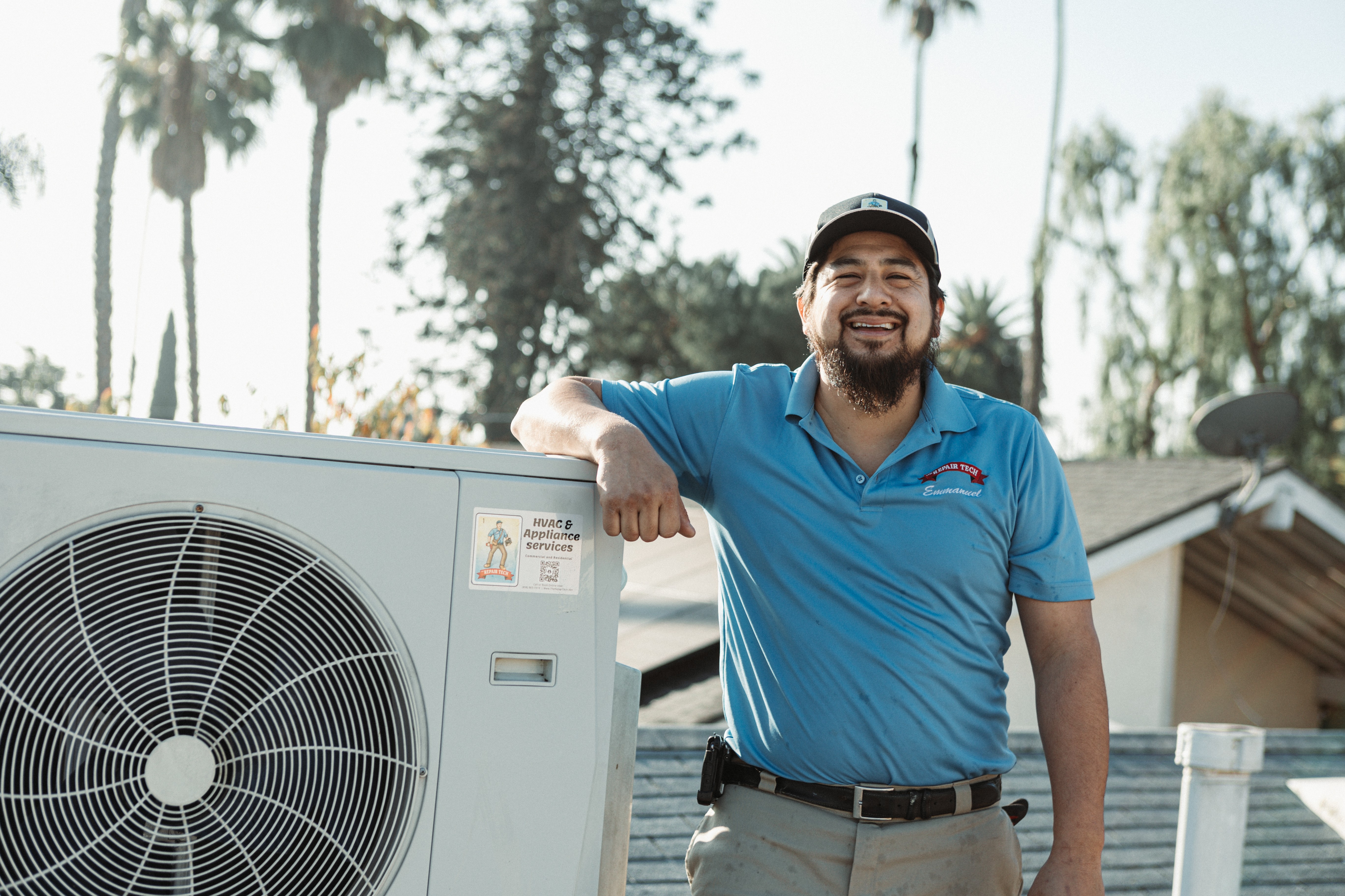 Man in blue polo shirt smiling outdoors, standing next to an air conditioning unit, with trees in background.