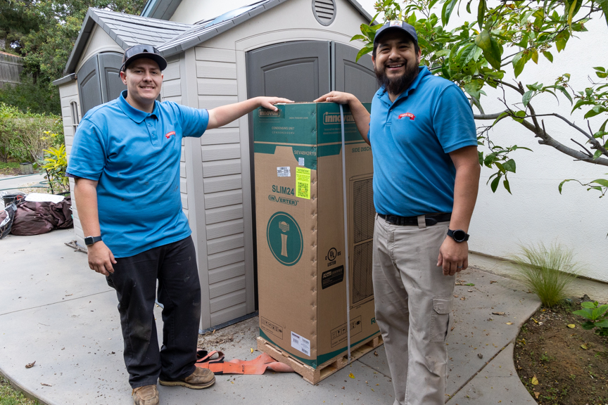 Two delivery workers in blue shirts stand outdoors next to a tall, boxed item, smiling and pointing at it.