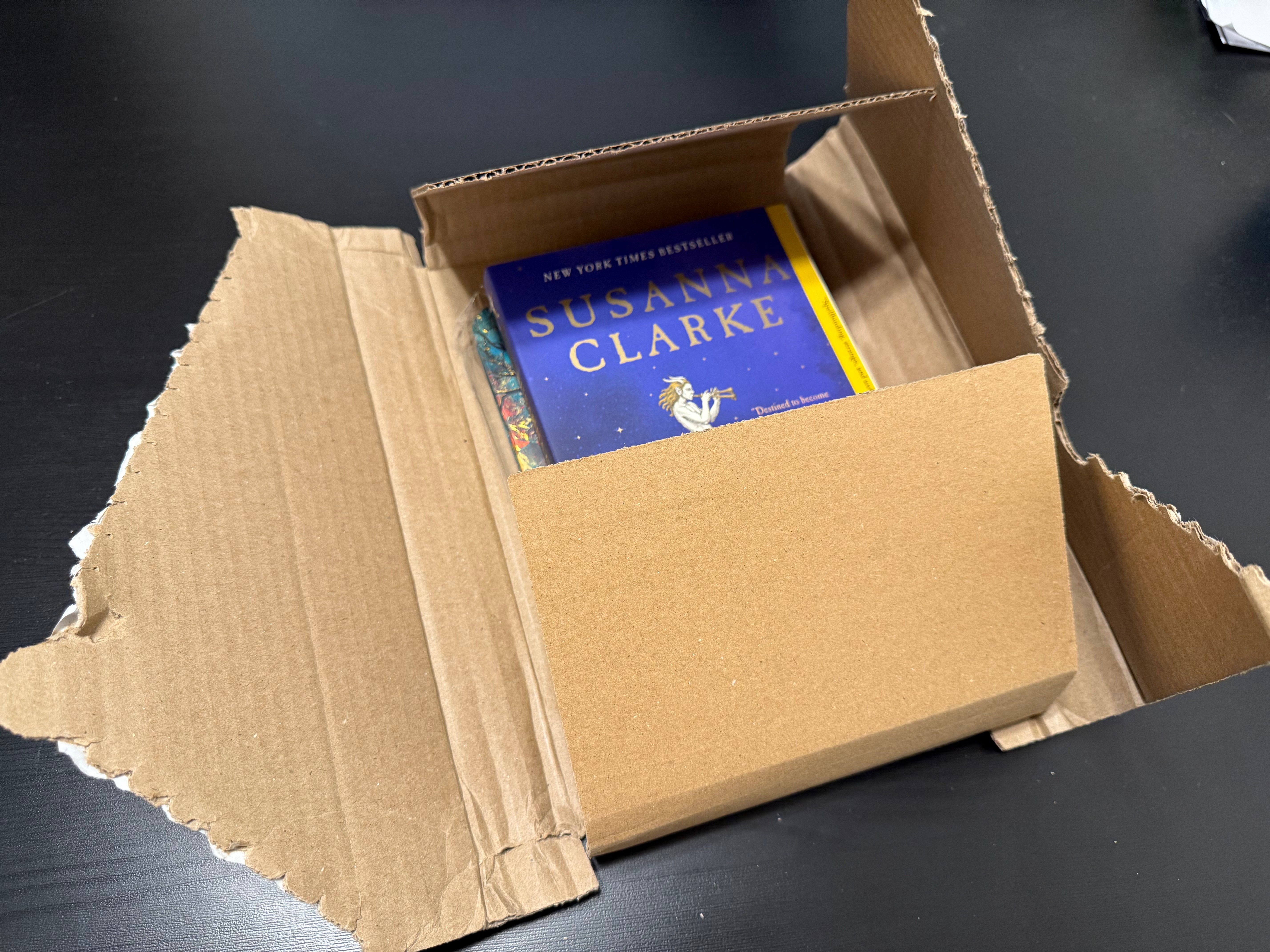 Open cardboard box with a book titled 'Susanna Clarke' inside, on a dark surface.