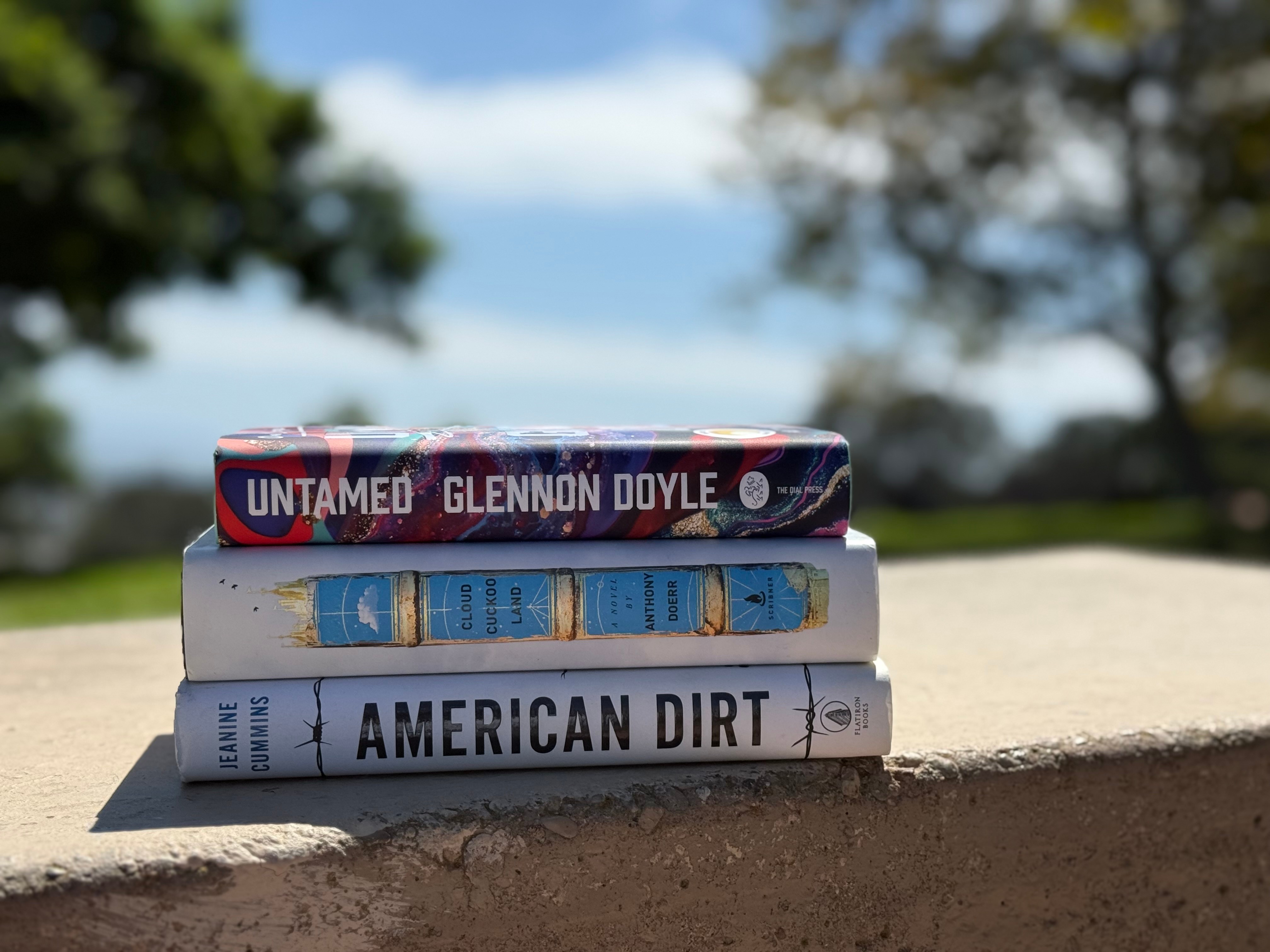 Three books stacked on a ledge outdoors with trees and sky in the background.