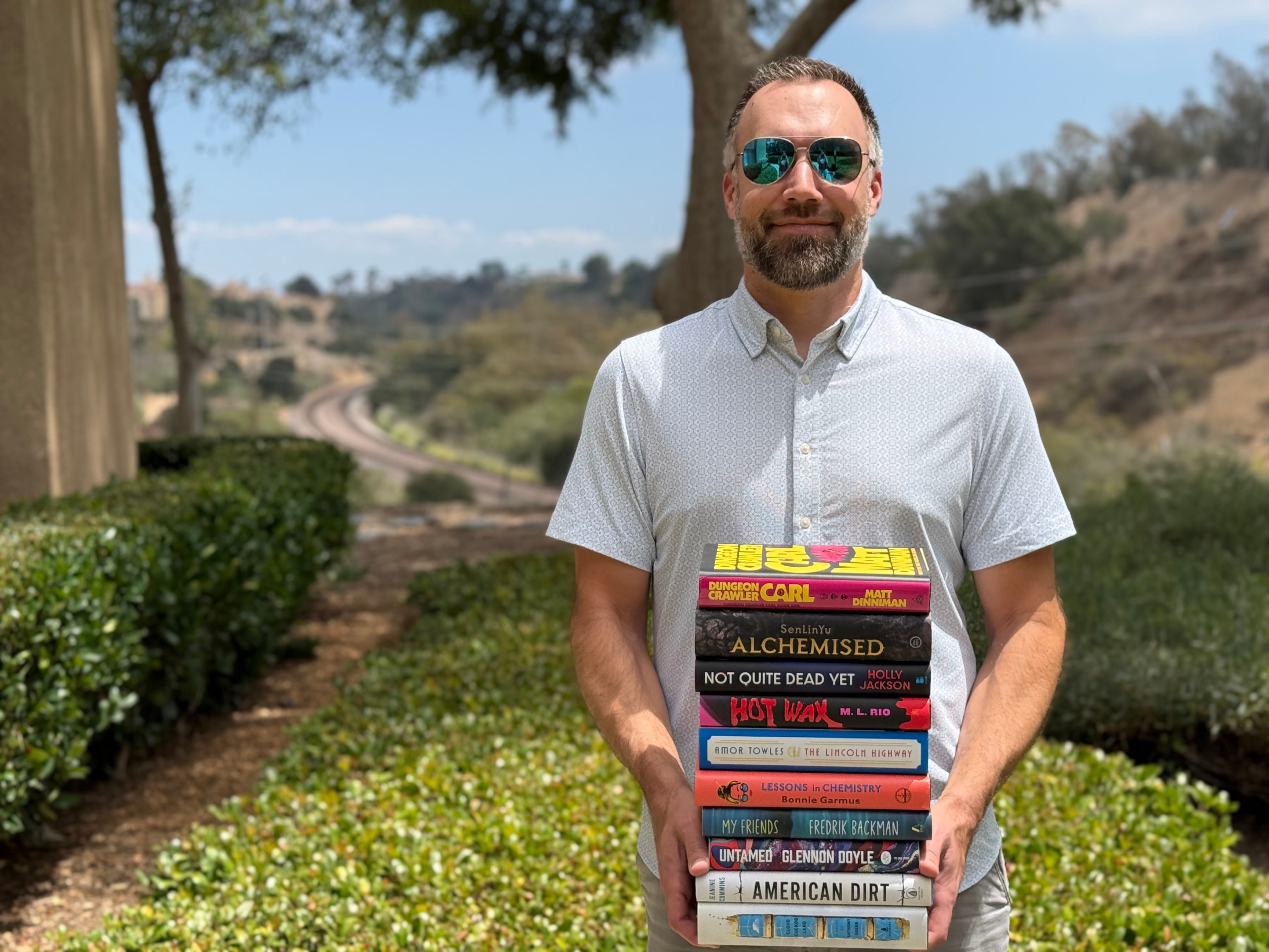 Man with sunglasses holding a stack of books outdoors, trees and hills in background.