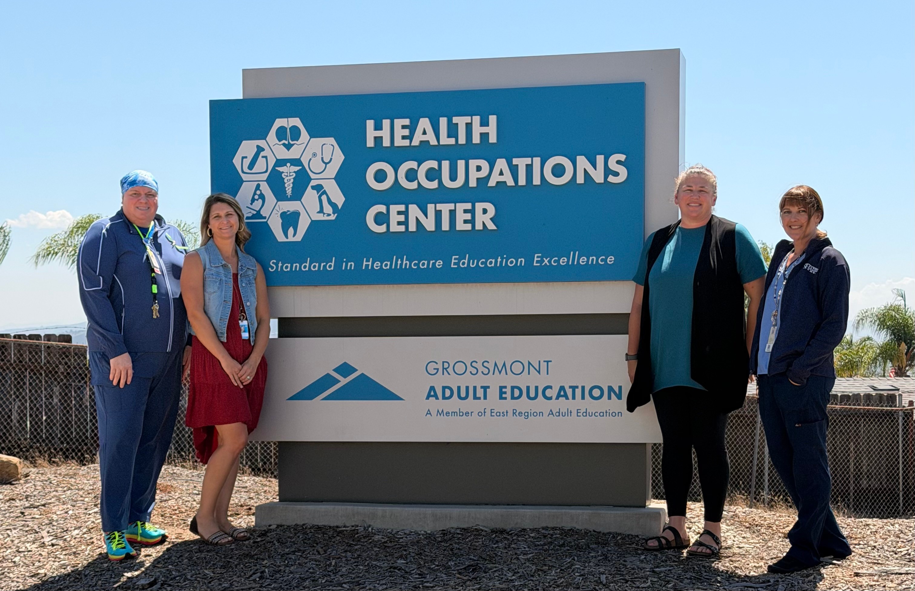 Four people standing outdoors next to a sign for a health occupations center, with a blue sky and clouds in the background.