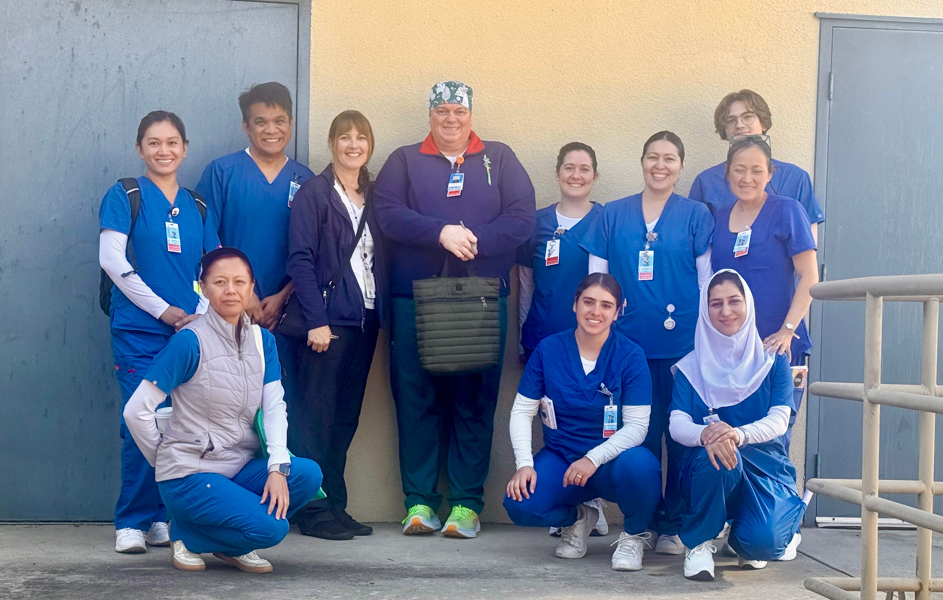 Group of ten healthcare workers in blue uniforms and one person in formal attire, standing outdoors in front of a building.