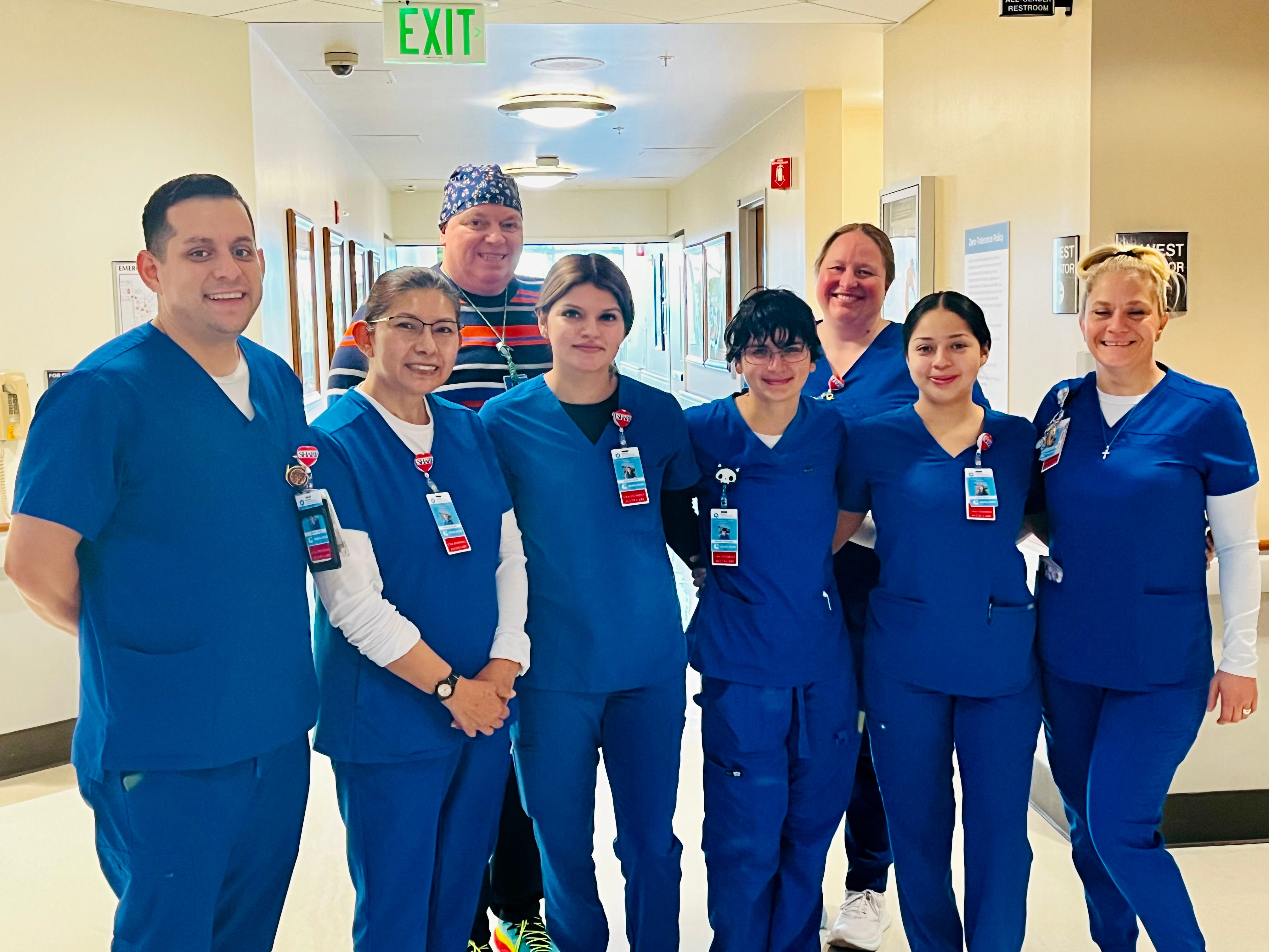 Group of seven healthcare professionals in blue scrubs standing in a hospital corridor, smiling at the camera.