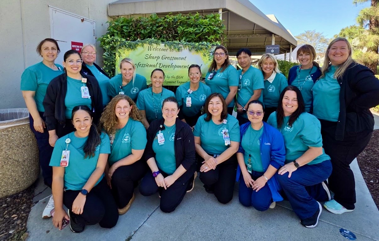 Group of 14 women in teal uniforms standing outdoors in front of a building with greenery and a sign, smiling.