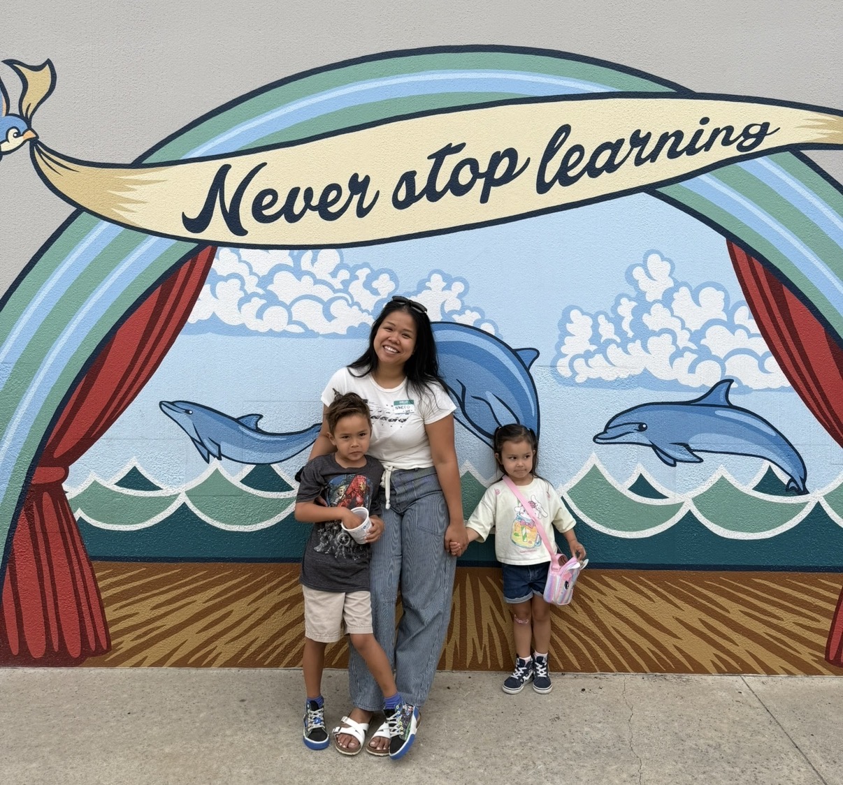 Three people standing in front of a colorful mural with dolphins and waves, and a banner reading 'Never stop learning.'