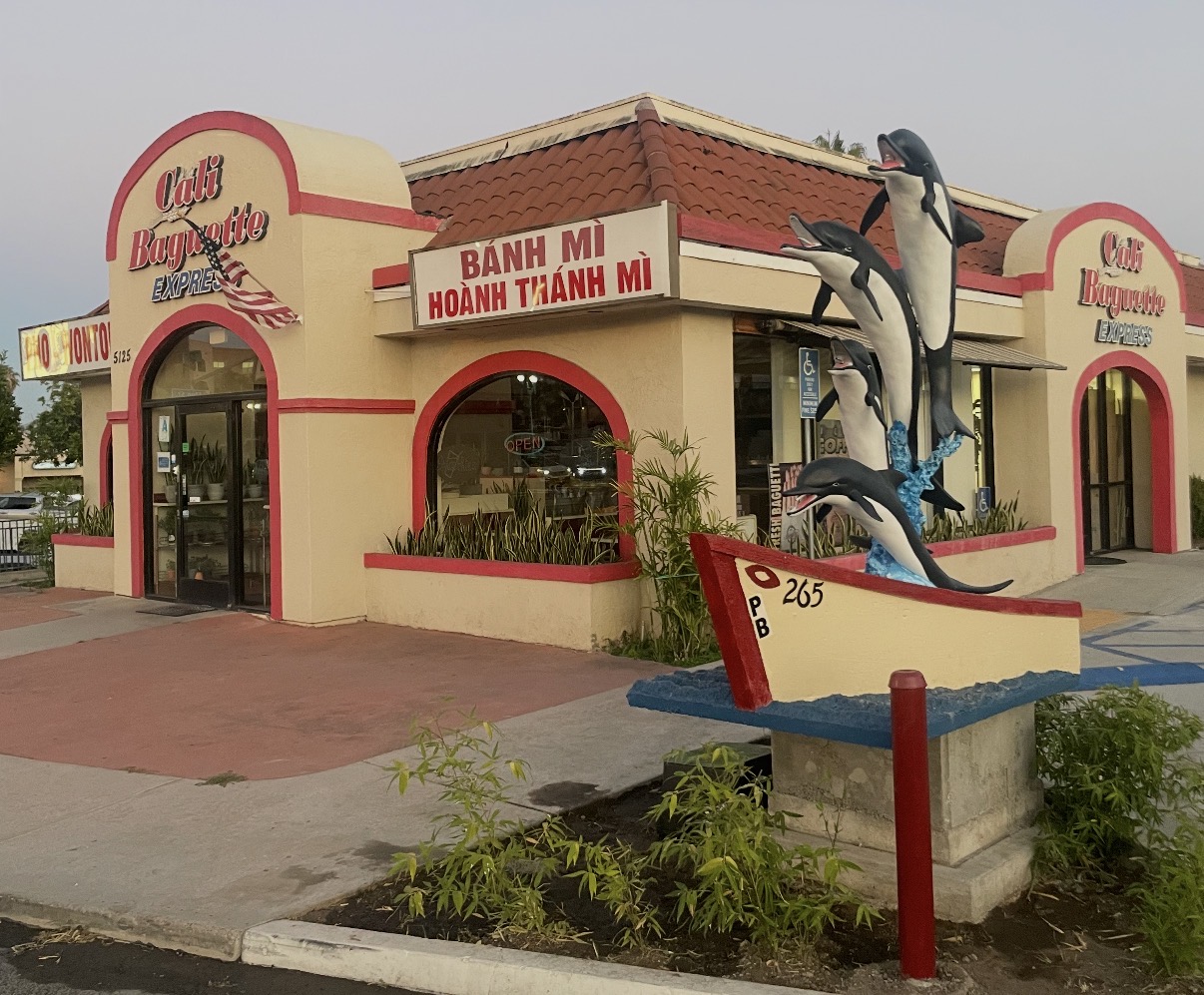 A restaurant building with dolphin sculptures outside, sign in Vietnamese, and a boat sculpture nearby, under a clear sky.